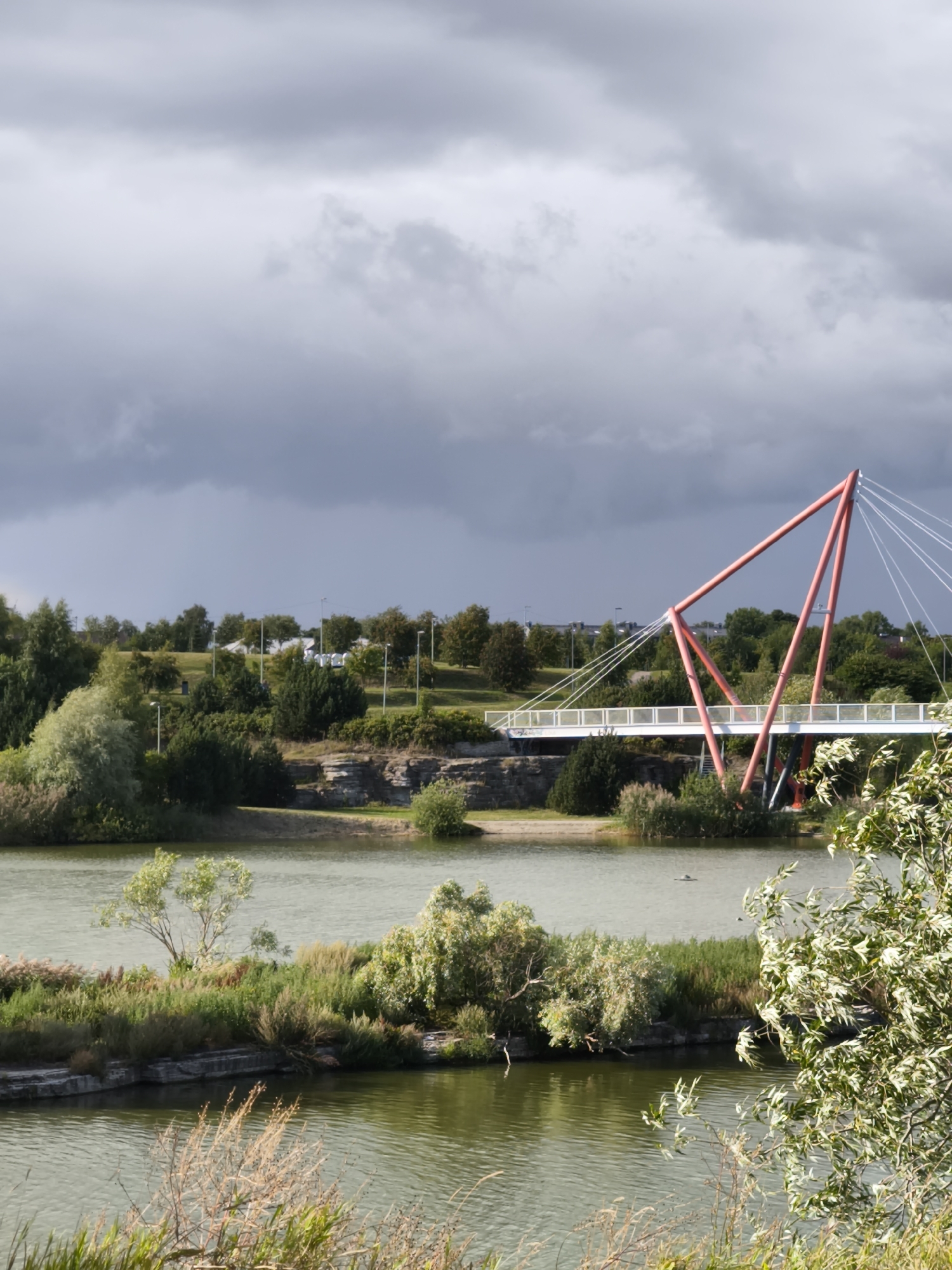 The image shows a modern cable-stayed bridge with a distinctive red triangular pylon spanning a body of water. The bridge has a white deck with railings. The surrounding landscape is lush and green, with trees, bushes, and reeds growing along the water's edge and on hills in the background. The sky is partly cloudy, with dramatic dark clouds suggesting impending rain. In the distance, on the hilltop, several portable toilets are visible, along with streetlights. This scene depicts the Pirita beach area in Tallinn, Estonia.