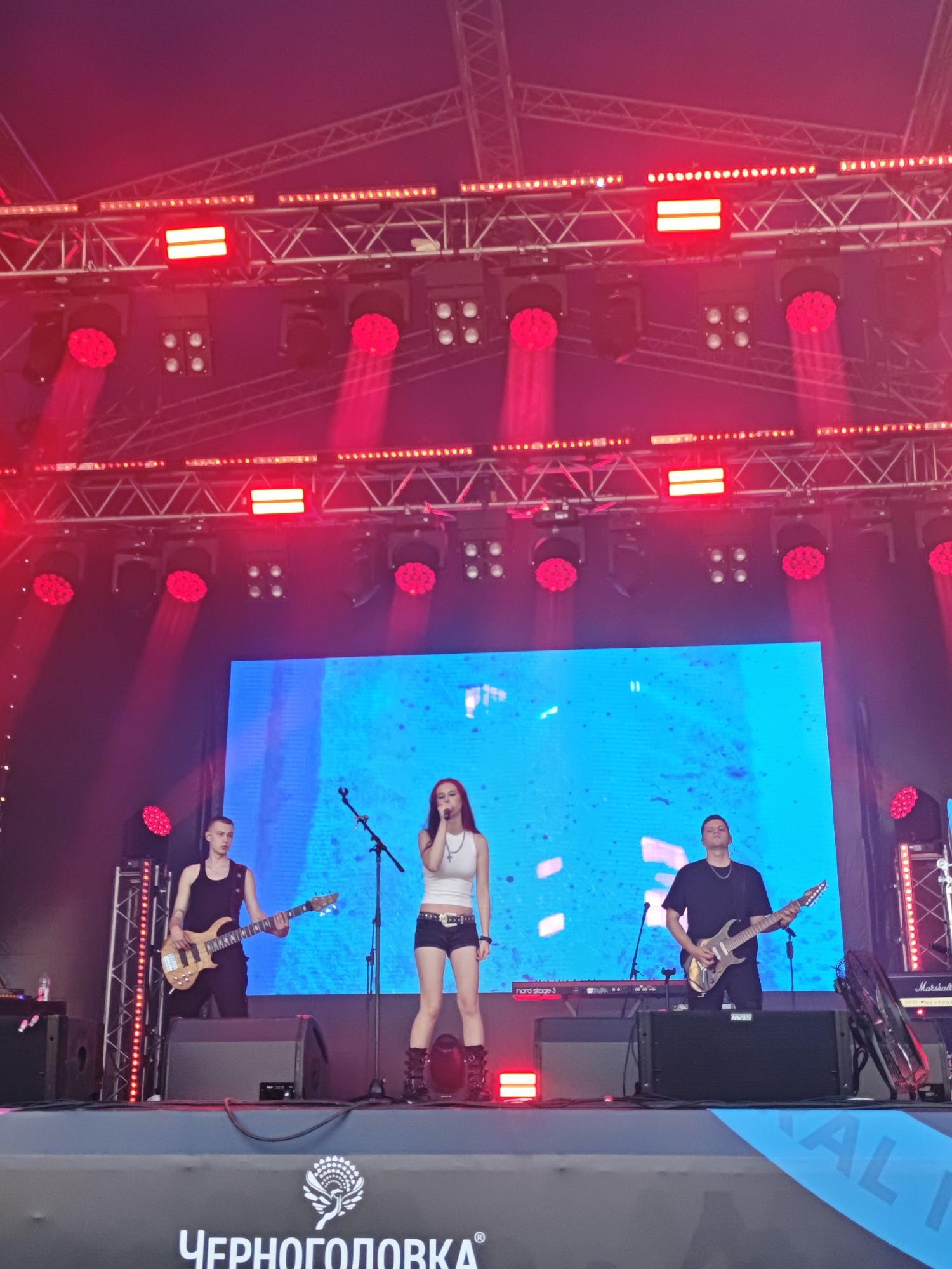 The image captures a live band performing on an outdoor stage. Three musicians are visible: a male bassist on the left, a female singer in the center, and a male guitarist on the right. The singer is wearing a white top and denim shorts, while the guitarists are dressed in black. Red stage lights dominate the scene, casting a vibrant glow. Behind the performers is a large blue LED screen displaying abstract patterns. The stage is equipped with various musical instruments and audio equipment, including amplifiers and keyboards. In the foreground, a banner with a logo and text, which appears to be in Russian, is partially visible. The overall impression is that of a lively concert event.