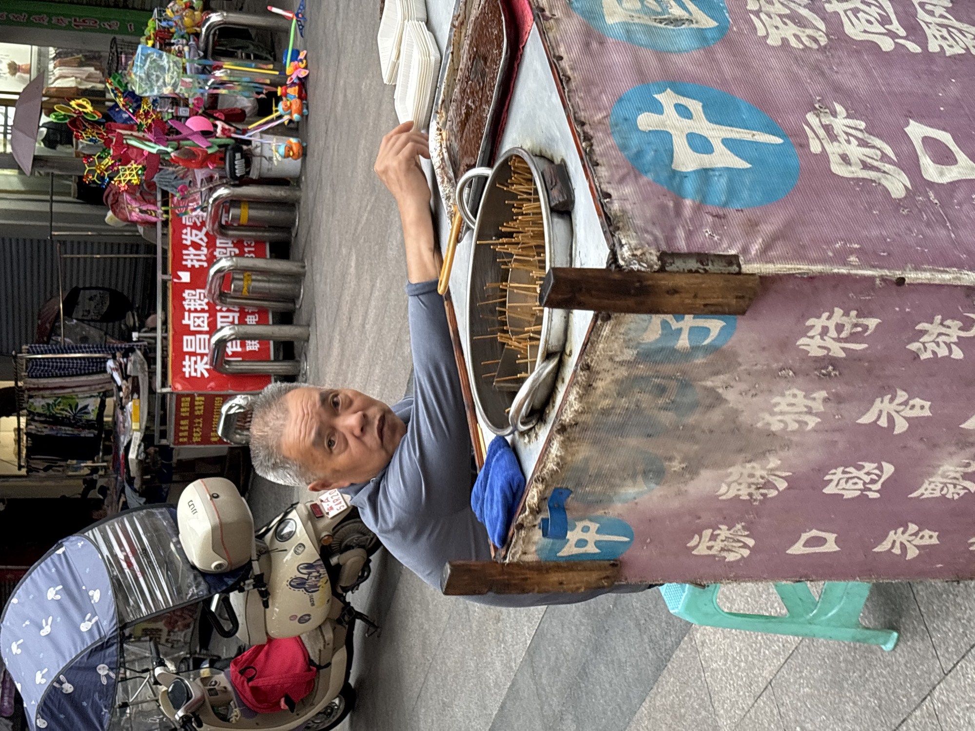An elderly Asian man with gray hair sits behind a food cart in Jiangjin, China. He is wearing a grey long-sleeved shirt and is looking towards the camera with a neutral expression. His right hand is resting on the cart, near a tray of food. The food cart is decorated with pink and purple fabric featuring white Chinese characters and blue circles with white characters. On the cart, there are metal containers with skewers of food and white paper containers. To the left of the man, a teal plastic stool is visible. Behind the cart, a scooter with a blue patterned umbrella is parked. The background shows a street scene with shops displaying clothing and toys, and metal barriers. The overall impression is that of a street vendor operating in a busy urban environment.