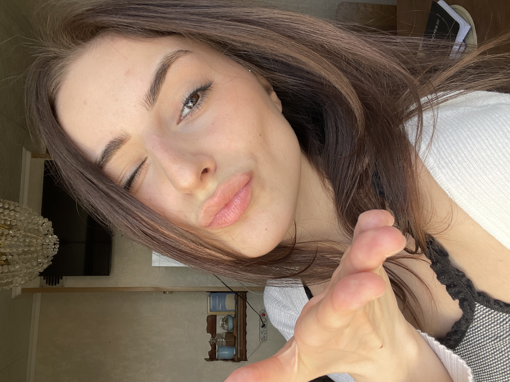 A young adult female with long, dark brown hair is centered in a close-up indoor portrait. She winks with her right eye and puckers her lips in a kissing gesture, directly facing the camera. Her left hand is extended towards the viewer, fingers slightly curled. She is dressed in a white ribbed long-sleeved top over a dark, possibly black, textured garment. The background features light-colored, textured walls and a white ceiling with molding. A crystal chandelier hangs from the ceiling in the upper-left. A flat-screen television is mounted on the wall in the upper-right quadrant. Below the television, a horizontal power outlet strip with multiple sockets and a red switch is visible. To the left of the power strip, a small, dark wooden wall-mounted shelf holds a light blue mug, two glass jars containing various substances, and a light blue rectangular box. A vertical wooden beam or door frame is present to the left of the television. A dark notebook labeled "NOTES" with additional papers rests on a light-colored surface on the lower-right side of the frame, partially obscured by the subject's hair. The scene is brightly illuminated. The location is an interior space within the city of Moscow, Russia.