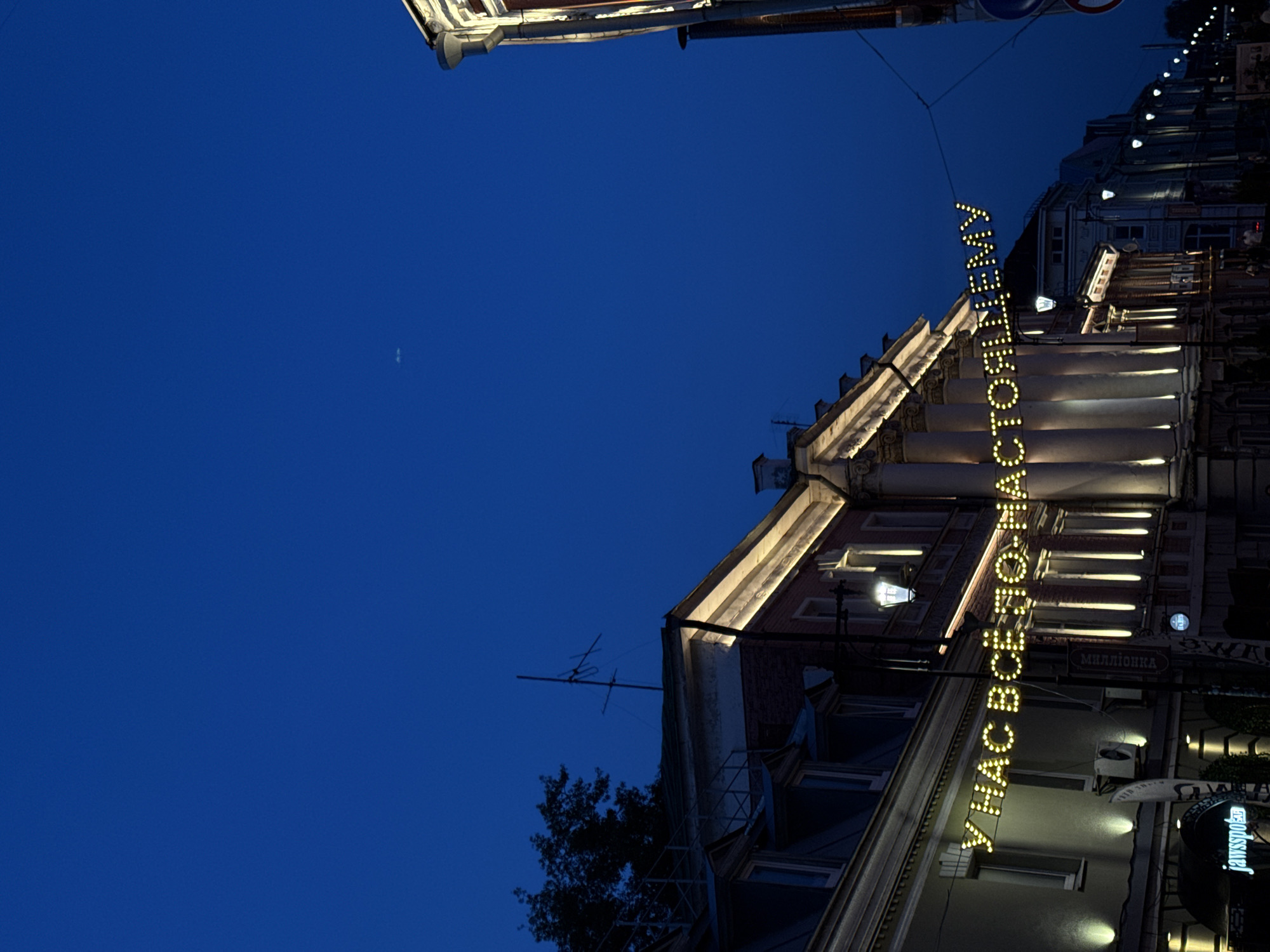 Captured at night, the photograph depicts a building in Nizhny Novgorod, Russia. The building's facade is illuminated with lights. A sign in Russian, vertically oriented, is also lit up. The sky is a deep blue. Adjacent buildings and streetlights are visible. Some foliage is present.