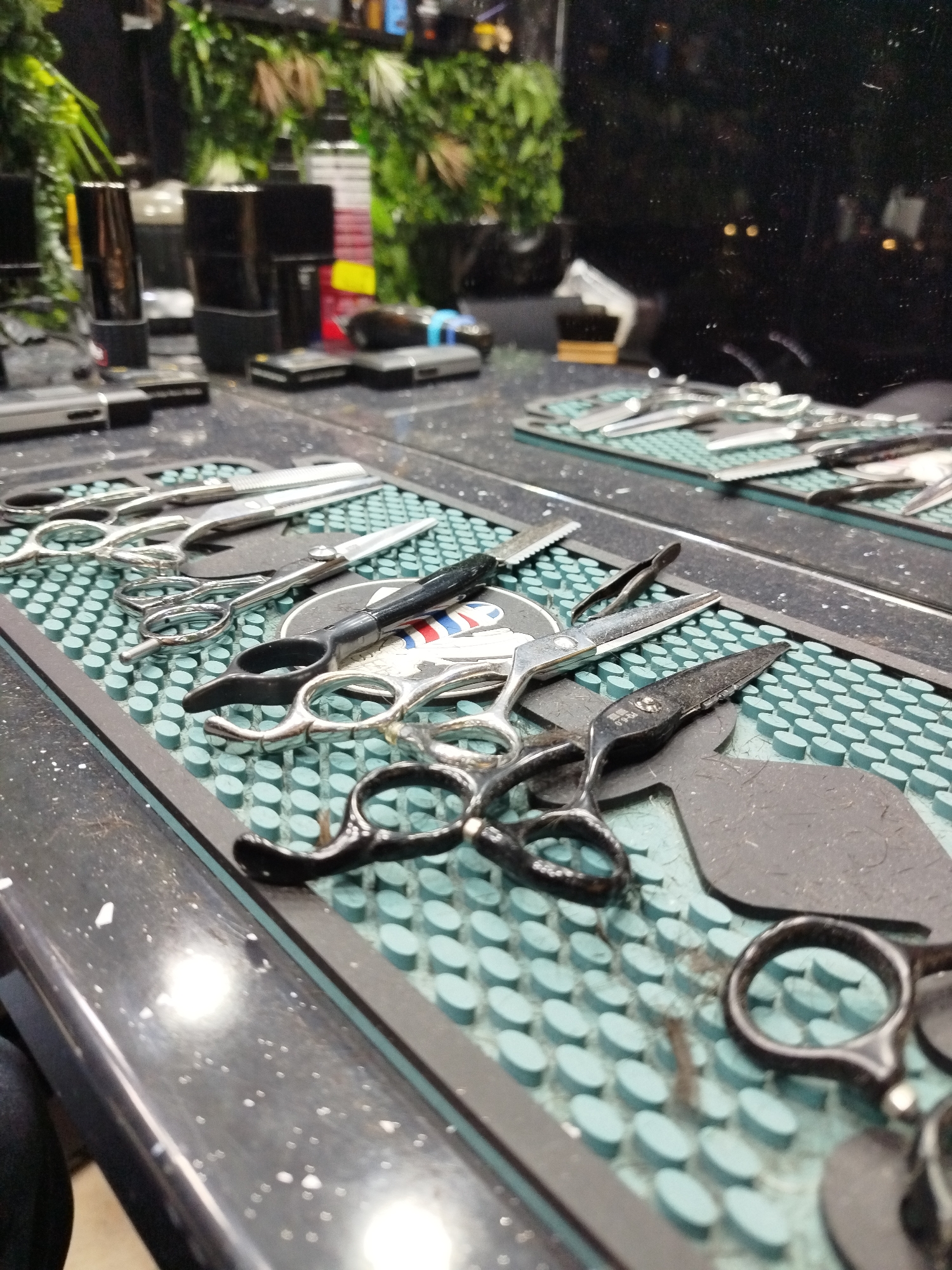 The image shows a barber's station. Several pairs of scissors are laid out on a light-blue, textured mat with small, raised circles, on a dark counter. Some scissors are silver-colored, while others have black handles. Behind the first row of scissors, there is another mat with more scissors visible. In the background, there are various barbering supplies such as clippers and hair products, as well as greenery. The scene seems to be indoors, possibly in a barber shop. The location is unknown, and it is impossible to confirm that it is in the city of Varna, Bulgaria. There are no people visible in the image.