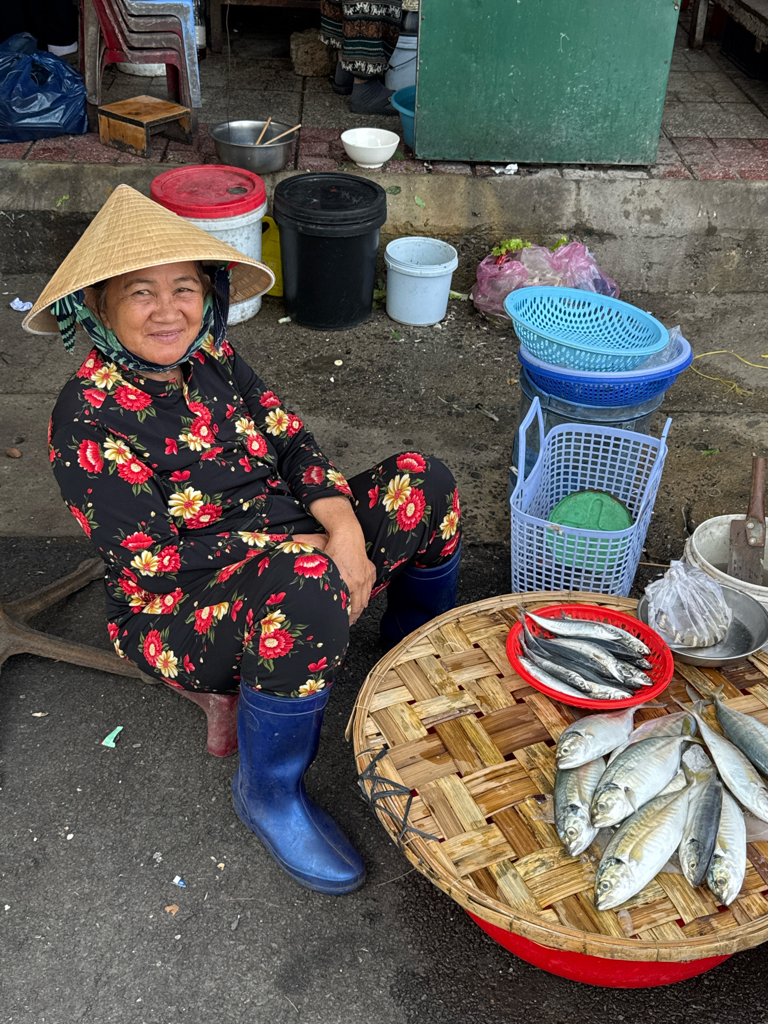 The image depicts a street scene in Nha Trang, Vietnam, where a woman wearing a conical hat and a floral patterned outfit is seated on the ground. She is looking at the camera and smiling. In front of her, on a round woven mat, are several fish. To her left, there are various containers, including buckets and baskets, and behind her, there are stacks of blue baskets. The ground is paved with asphalt and there is some debris scattered around. The overall impression is of a market stall or a vendor selling fish on the street.