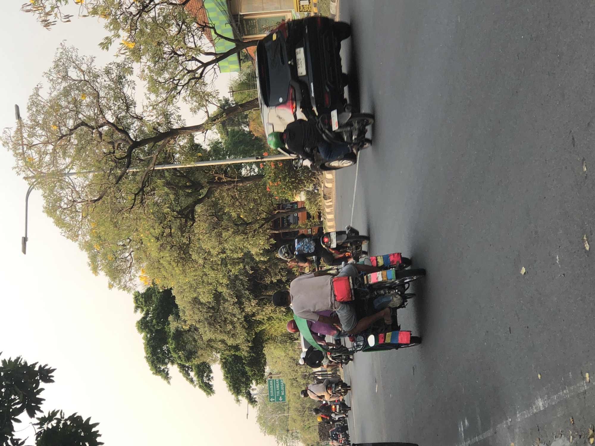 The image depicts a street scene in Kludan, Indonesia, with multiple vehicles and individuals navigating a paved road. In the foreground, a three-wheeled vehicle, possibly a modified motorcycle or auto-rickshaw, is being ridden by at least two people. The vehicle is adorned with colorful stripes on its rear. Other motorcycles, some with passengers, are visible in the mid-ground, suggesting moderate traffic.
Further in the background, a black SUV is on the road. The environment includes trees with green foliage and some yellow flowers, indicating a potentially tropical or sub-tropical setting. A street sign is also visible, pointing towards "SURABAYA" and "TULANGAN - MOJOSARI," providing a specific geographical context within Indonesia. The sky is overcast with a light, hazy quality, suggesting it might be dawn, dusk, or a cloudy day. The overall impression is of a typical urban or suburban street with active transportation.