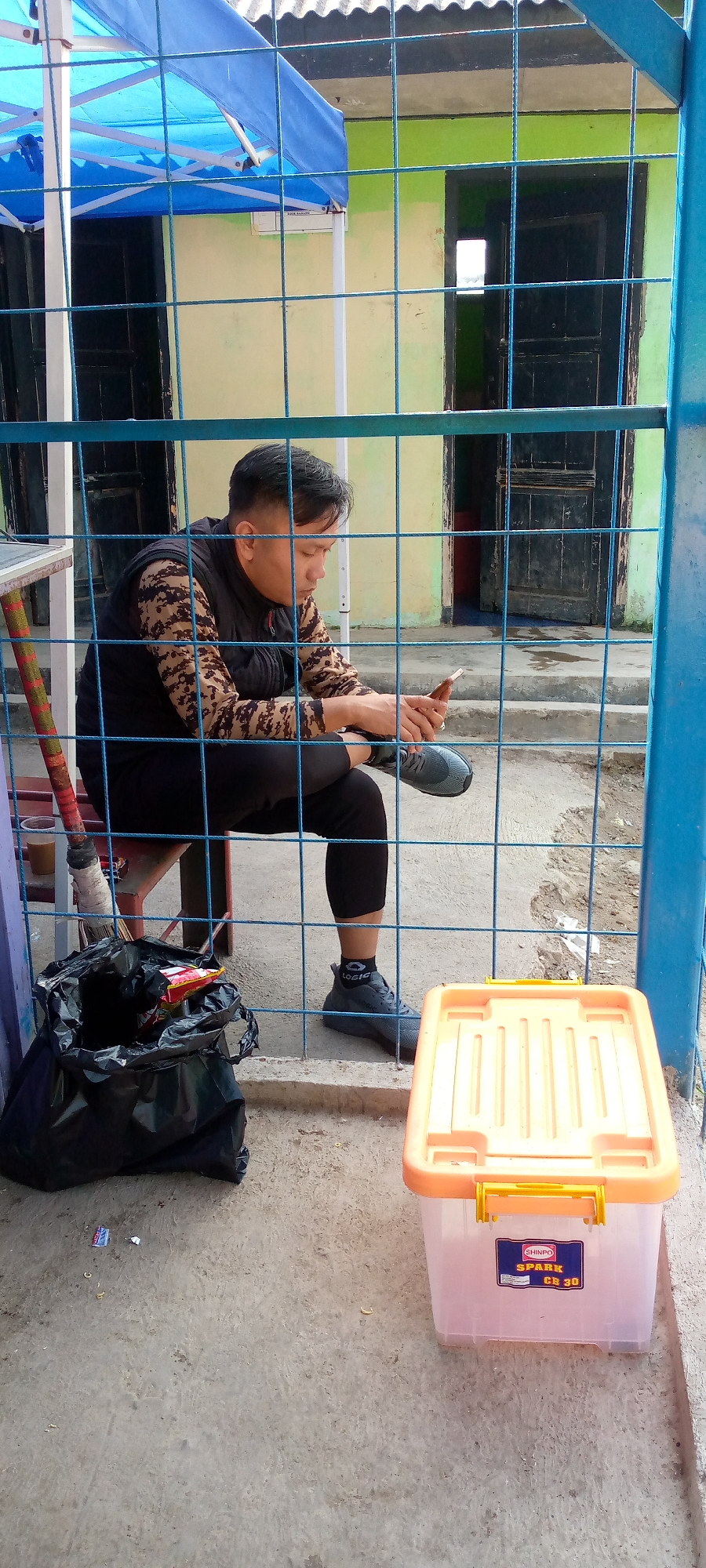 A man is sitting on a red chair behind a blue metal fence. He is wearing a camouflage long-sleeved shirt and a black vest. He is holding a smartphone in his right hand and looking down at it. His left leg is crossed over his right leg, and he is wearing grey athletic shoes. To his left is a black trash bag filled with items, and to his right is an orange and clear plastic storage container. The background consists of a light yellow wall with a dark door and a blue canopy overhead. The ground is concrete. The scene appears to be outdoors, possibly in a yard or public space in Subang, Indonesia.