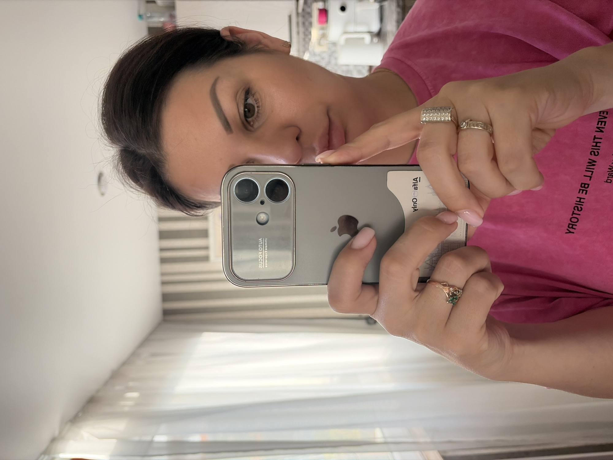A woman with dark hair is taking a selfie in a room with natural light coming from a window with sheer curtains. She is wearing a bright pink t-shirt with text on it that is partially visible. The woman is holding a silver smartphone in her left hand, which is reflecting the room. She is wearing two rings on her right hand and a ring with a green stone on her left hand. The background shows a wall with striped wallpaper and a kitchen counter with appliances.