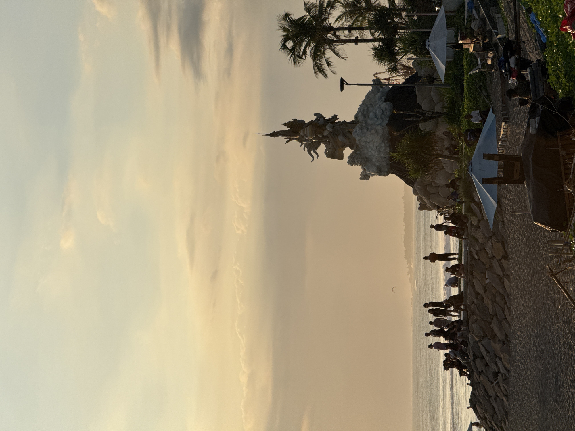 The image captures a bustling scene at sunset, likely in or around North Kuta, Indonesia, judging by the tropical vegetation and seaside location.
In the center is a large, ornate statue depicting multiple figures. It's positioned on top of a rocky structure with dense vegetation at its base.
A crowd of people is visible along the rocky shoreline, many of whom seem to be observing the sunset. Some are standing on the elevated, man-made rocks near the beach.
Several structures, possibly small stalls or temporary shelters, are on the edge of the scene, indicating a public or commercial space.
The sky is dominated by hues of yellow and orange. The overall lighting indicates the photo was taken during the evening.