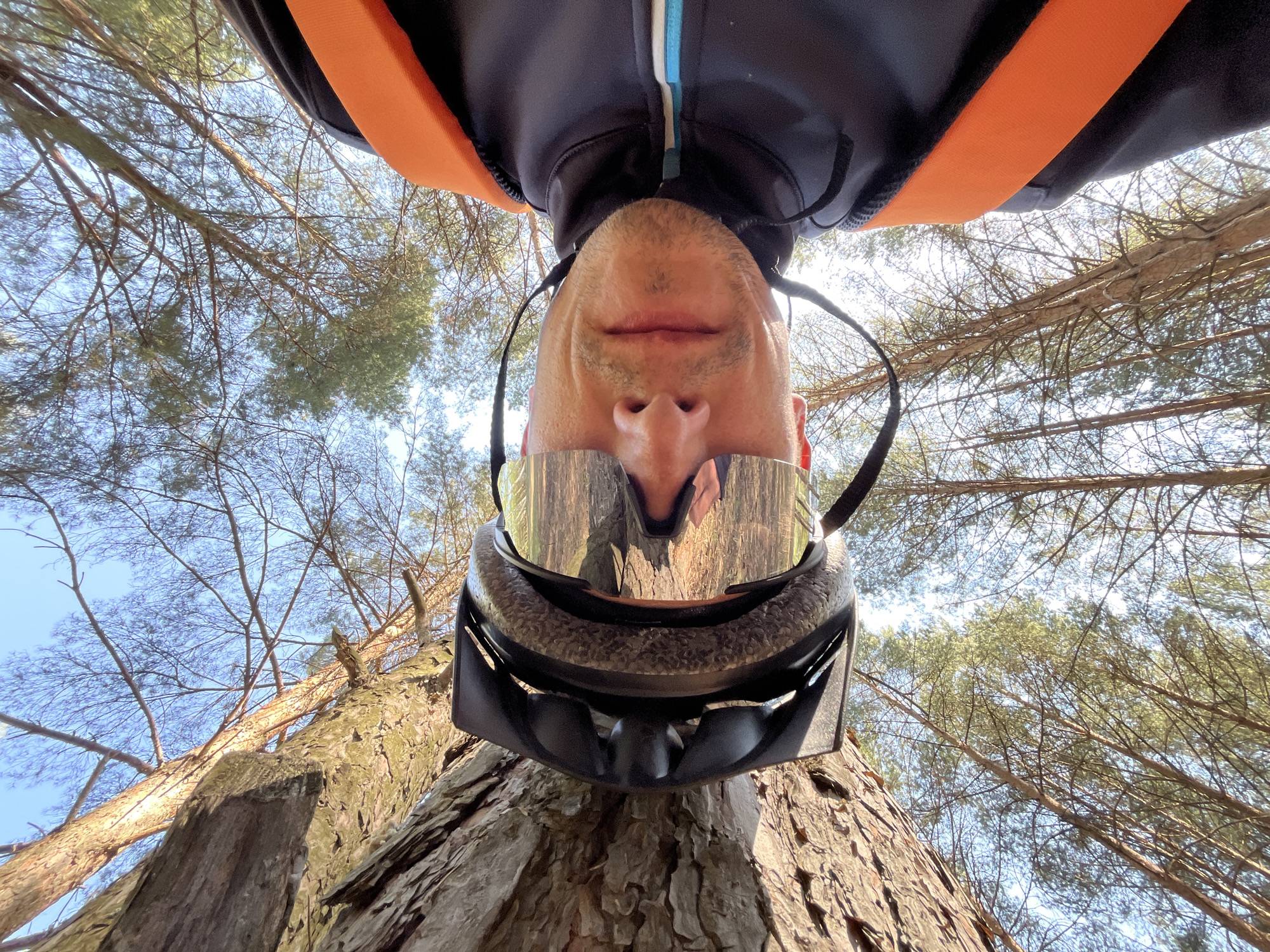 A low-angle perspective photograph depicts a man wearing a helmet and reflective sunglasses, seemingly lying on his back and looking upward. The trees frame the shot, and the sky is visible through the branches. The location is Chaikovsky, Russia. The man's face is positioned at the top center of the frame, while the helmet and sunglasses occupy the middle ground. The trees and sky form the background. The man appears to be outdoors.