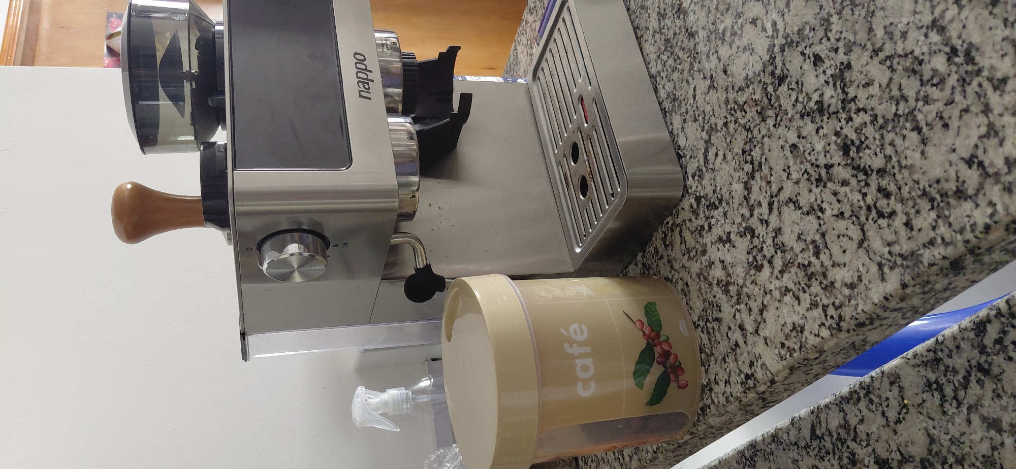 The image shows a close-up of a kitchen counter in Encarnacion, Paraguay, featuring a stainless steel espresso machine with a wooden tamper and a clear plastic coffee canister labeled "café". The counter is made of granite with a speckled pattern of beige, black, and grey. To the left of the coffee canister, a clear spray bottle with a white nozzle is visible. The espresso machine has a control dial on the left, a portafilter holder, and a drip tray. The brand name "nappo" is visible on the front of the machine. The background includes a white wall and a wooden doorframe. The lighting suggests an indoor setting, likely a home kitchen.