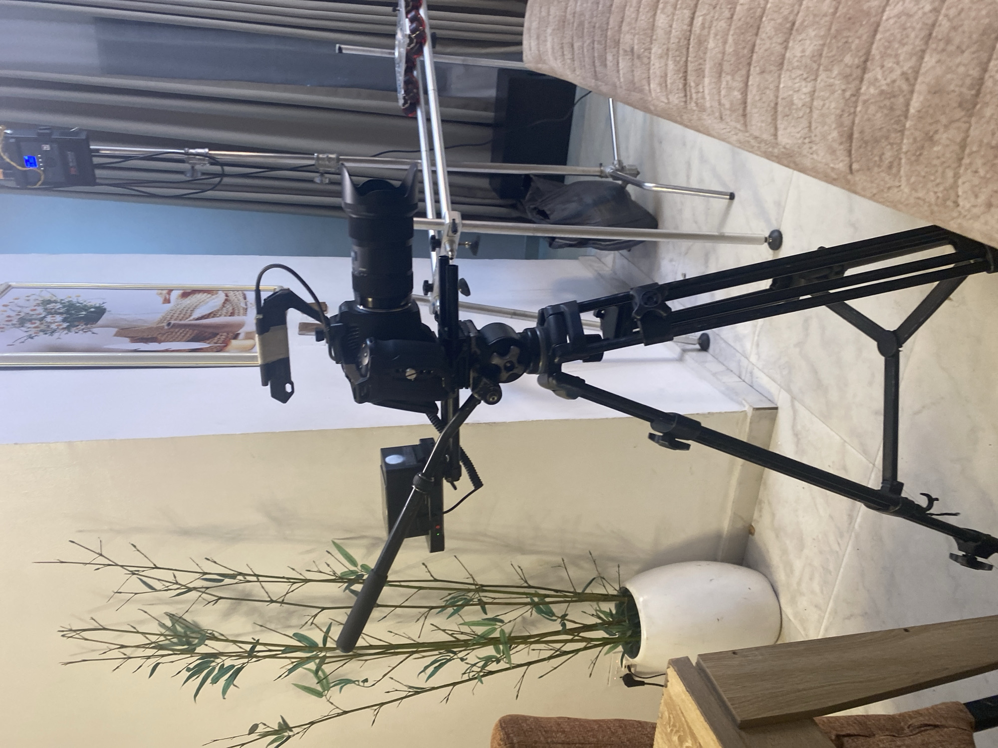 The image shows a professional camera setup on a tripod in an indoor setting. The camera is a black DSLR or mirrorless type with a large lens attached, and it's mounted on a heavy-duty tripod. The tripod has black legs and is positioned on a light-colored tile floor. Behind the camera, there's a white wall with a framed picture and a plant in a white pot. In the background, there are curtains, a light stand, and part of a couch.