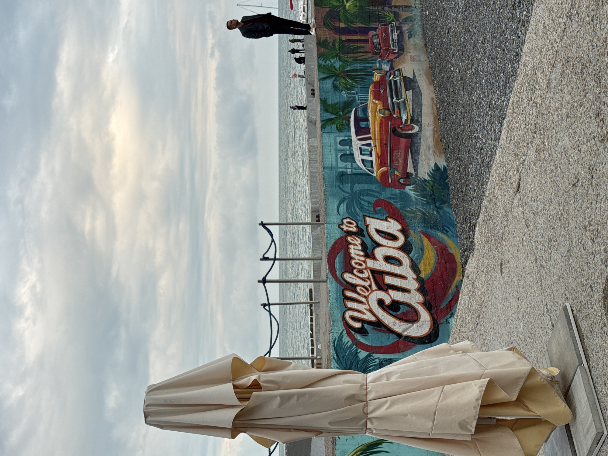 The image depicts a coastal scene, likely a waterfront area in Sochi, Russia. A mural featuring the phrase "Welcome to Cuba," along with classic Cuban cars and palm trees, adorns a concrete wall beside a pebbled area. A closed, tan-colored umbrella is visible in the foreground. In the background, there is a view of the sea, with a metal railing separating the waterfront from the walking area. Several people can be seen walking along the promenade, and one man is leaning against the wall near the mural. The sky is overcast with cloudy conditions.