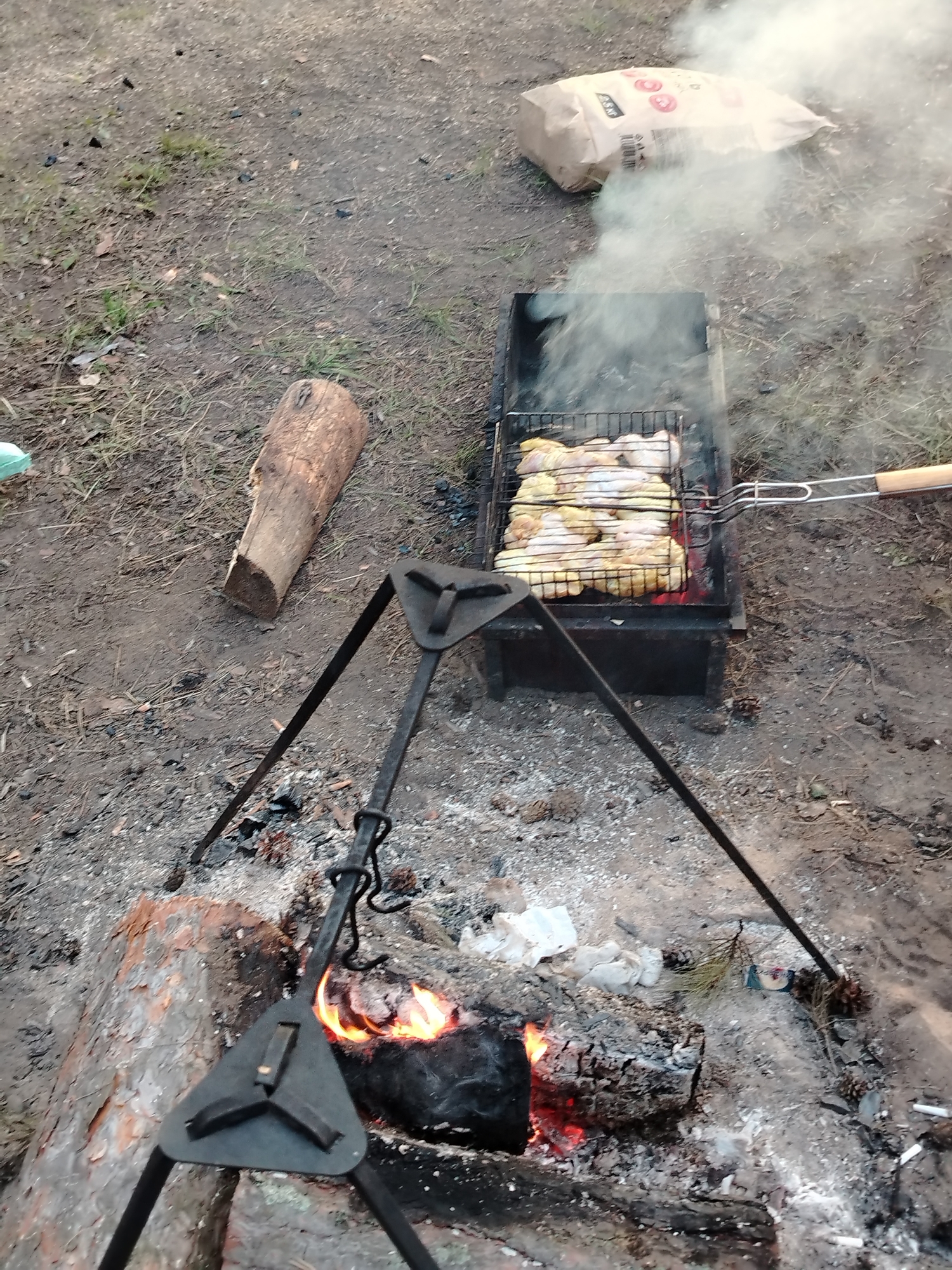 The image depicts an outdoor cooking scene. In the foreground, a fire burns in a makeshift fire pit made of logs. A metal tripod stands over the fire pit. To the right, a grill is placed over a metal brazier, cooking what appears to be chicken. Smoke rises from the grill. In the background, a paper bag, possibly containing charcoal, sits on the ground. The surrounding environment consists of dirt and scattered debris. The scene is likely located in the city of Chaikovsky, Russia, based on the provided context.