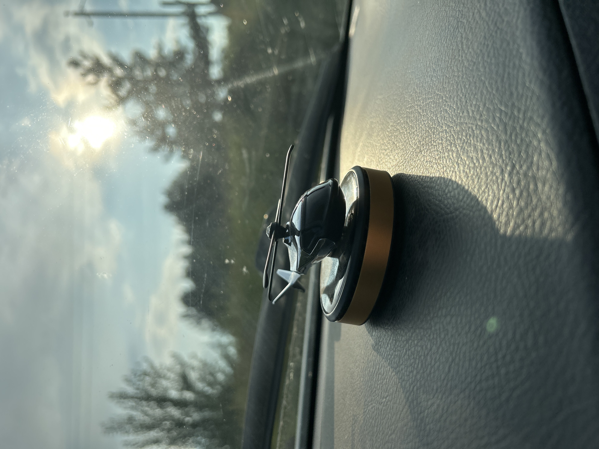 A black and gold toy helicopter sits on a dark gray, textured dashboard inside a vehicle. The helicopter has spinning rotors and is positioned to the left of the frame, with its base casting a shadow. The dashboard appears to be made of leather or a similar material. Through the windshield, which shows some dirt or water spots, a blurry outdoor scene is visible. The sun is shining brightly in the upper right portion of the frame, partially obscured by trees and clouds. The overall lighting suggests it is daytime, likely late afternoon due to the angle of the sun. No people are visible in the image. The specific location within Noviy, Russia, cannot be determined from the visual information provided.