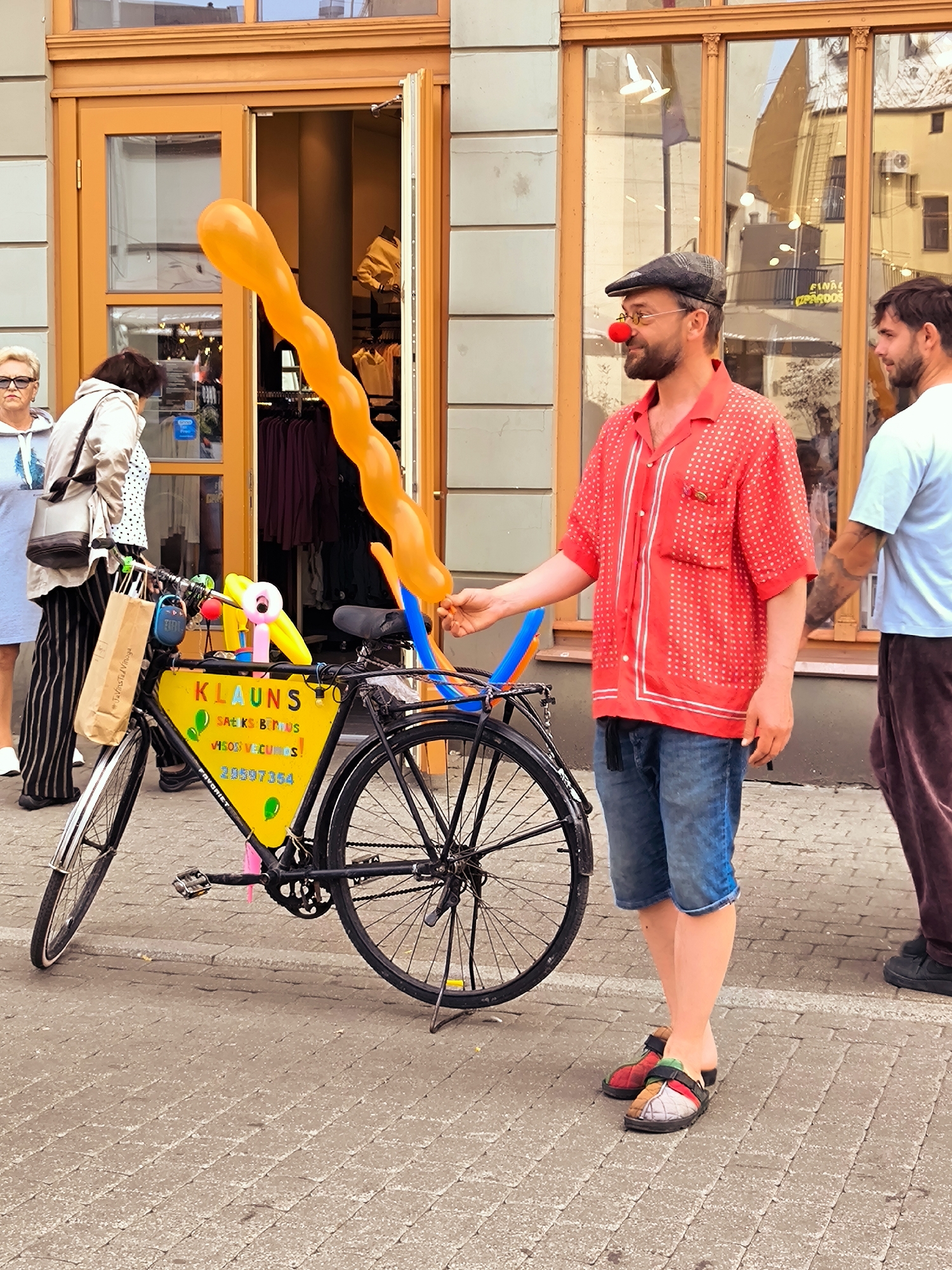 In Riga, Latvia, on a cobblestone street outside a building with an orange facade and large glass windows, a clown wearing a red polka-dot shirt, blue denim shorts, a flat cap, and a red clown nose stands by a bicycle. The bicycle is adorned with a yellow triangular sign that reads "KLAUNS" (Clown in Latvian), along with other text and green balloon graphics. Attached to the bicycle are several colorful balloons, including a long, orange, wavy balloon. A second man in a light blue t-shirt and maroon pants stands to the right, facing the clown. Two women are visible in the background, one on the left wearing a light blue top and the other in a patterned shirt. The scene appears to be a street performance or promotion.