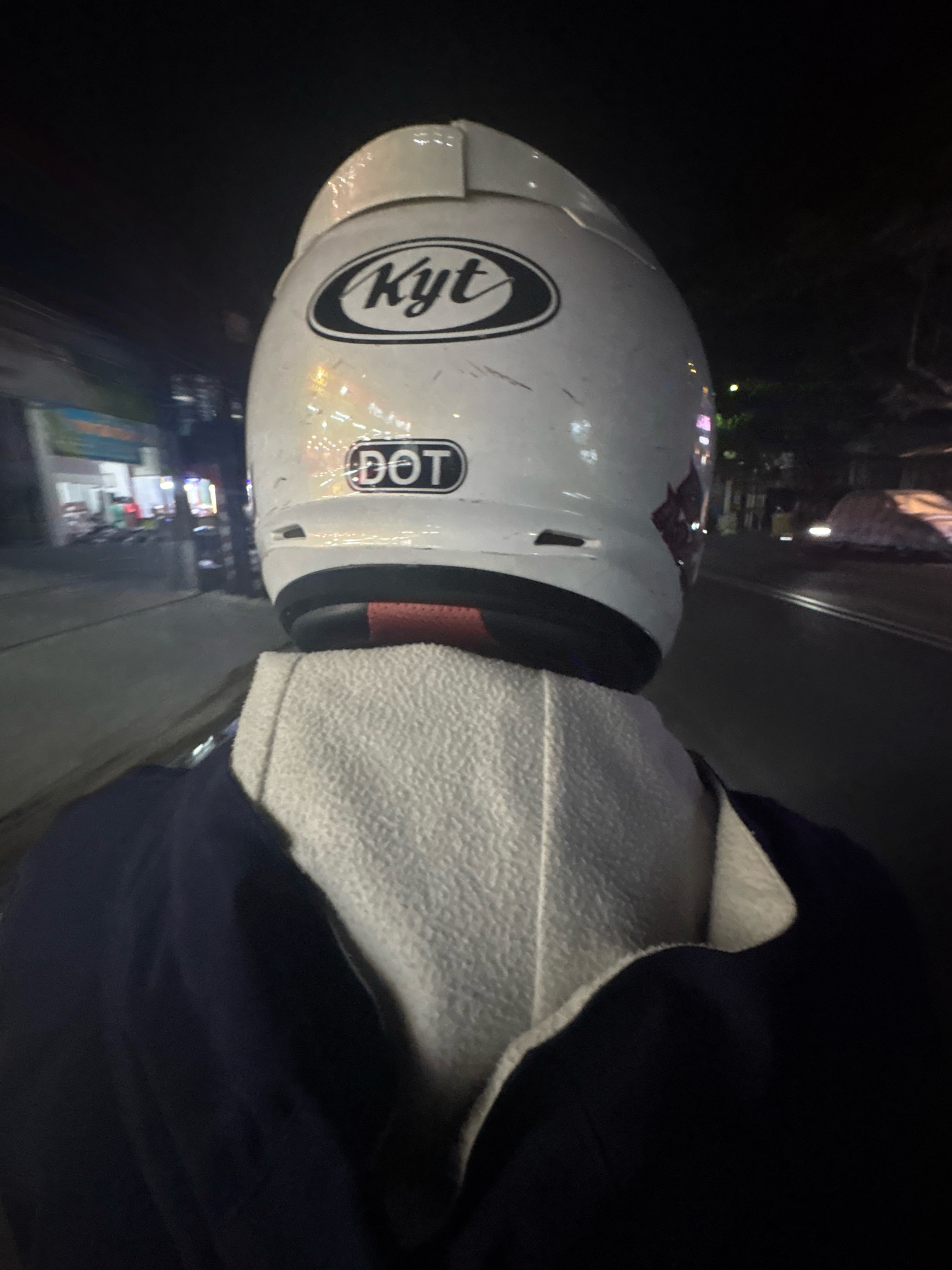 A low-light, rear-facing, first-person perspective shot captures a person wearing a white KYT brand helmet with a DOT safety certification sticker, riding a motorcycle at night. The rider is also wearing a dark blue hooded jacket over a light gray textured hooded shirt. The background shows a street scene with buildings and streetlights on the left, and a dark road with faint reflections and parked vehicles on the right. The overall impression is of a night ride through a moderately lit urban environment, likely in Bandung City, Indonesia, given the prominent KYT brand which is popular in Southeast Asia.