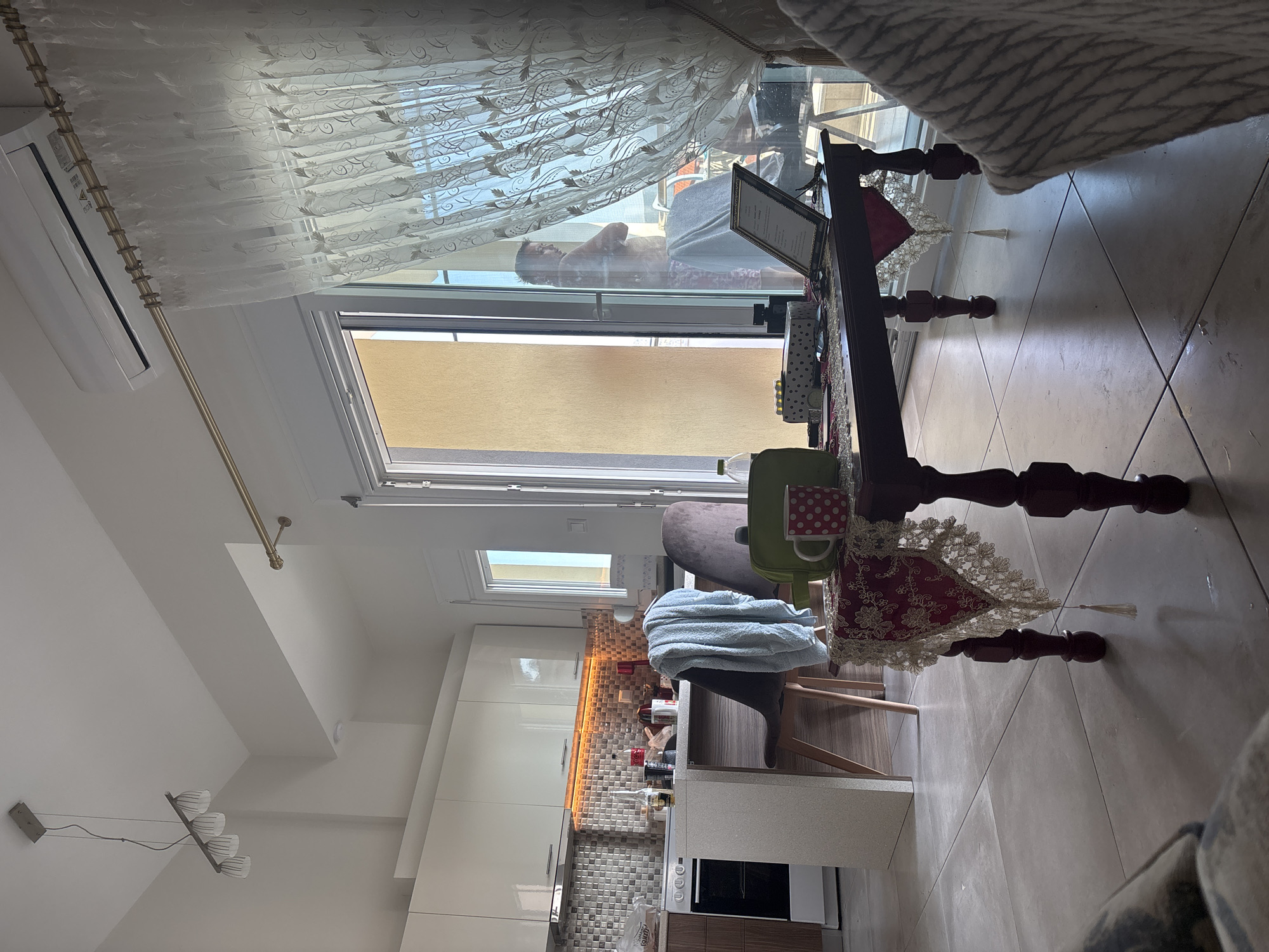 The image depicts an indoor scene, likely a living area or kitchen, in an apartment located in Νέα Ηρακλείτσα, Greece. In the background, a kitchen with white cabinets and a tiled backsplash is visible. A counter with a stove and oven is also present. In the foreground, a dark wooden table is set with decorative items, including a red and white polka dot mug and a framed document. A person, partially obscured by a sheer curtain, stands near a sliding glass door that leads to an outdoor space. The room is well-lit, with natural light streaming in from the sliding door and artificial light fixtures on the ceiling. The overall impression is that of a modern, lived-in apartment.