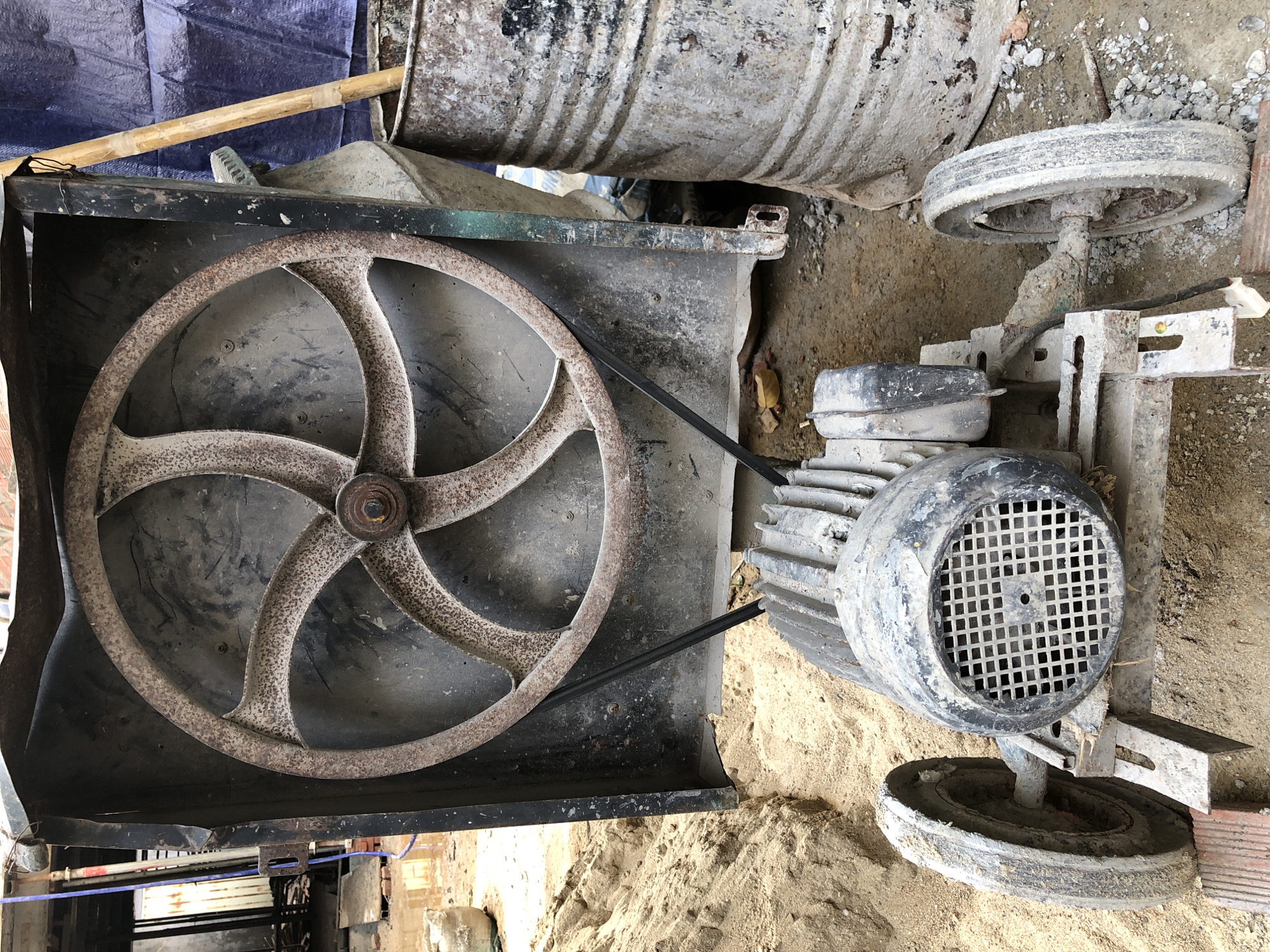 The image depicts a rusty, old industrial fan or mixer, likely abandoned at a construction site in Da Nang, Vietnam. The fan, with its five-spoked metal wheel, is covered in dust and rust, suggesting prolonged exposure to the elements. A black belt connects the fan wheel to a central motor, which is also visibly aged and grimy. The machine sits on a makeshift trolley with two solid rubber wheels, indicating it was once mobile.
The surrounding environment is a construction zone. Piles of sand are visible, a common material in such sites. In the background, a large, ribbed metal barrel stands, also showing signs of wear and tear. A blue tarp and wooden poles suggest ongoing construction or work activity, possibly for shelter or support. The ground is a mixture of dirt, sand, and debris, typical of an unfinished building area. The overall impression is one of disuse and decay within an active or recently active construction space in Da Nang.