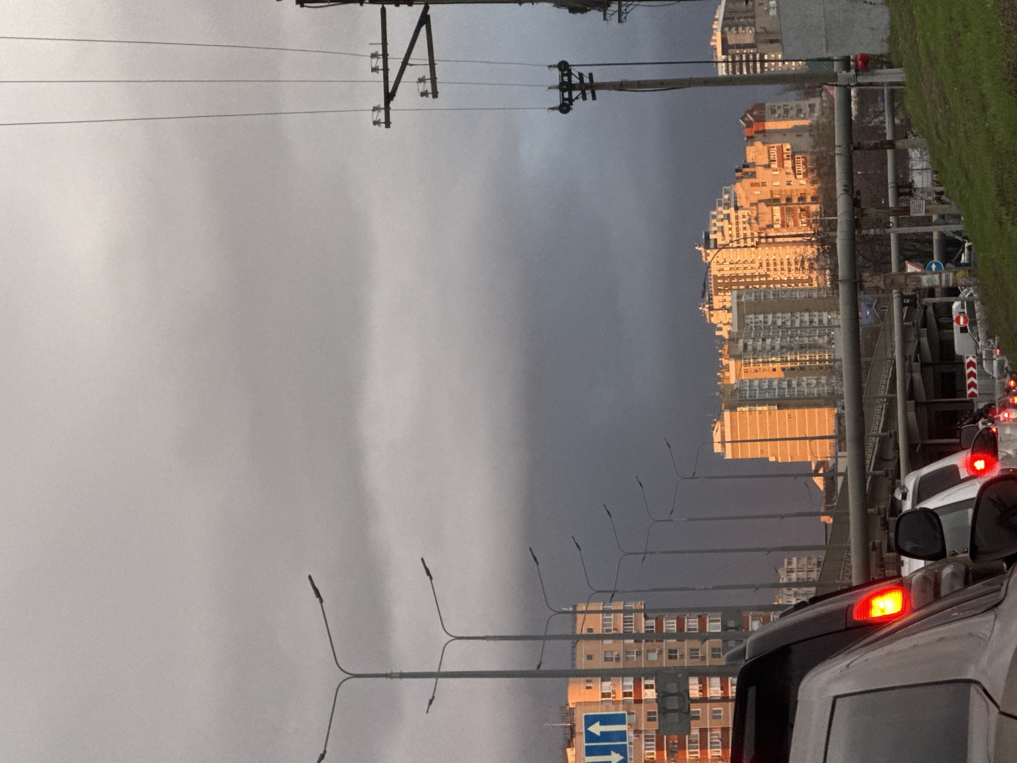 The image captures a street scene in an urban setting, most likely during twilight or a cloudy day. The lower half of the image shows traffic congestion, with cars positioned close to each other, some with their brake lights illuminated.
In the background, there are densely packed buildings. The buildings appear to be residential or commercial, with a mix of older and more modern architecture. Street lights stretch along the roadside.
The sky is overcast. Power lines are visible.