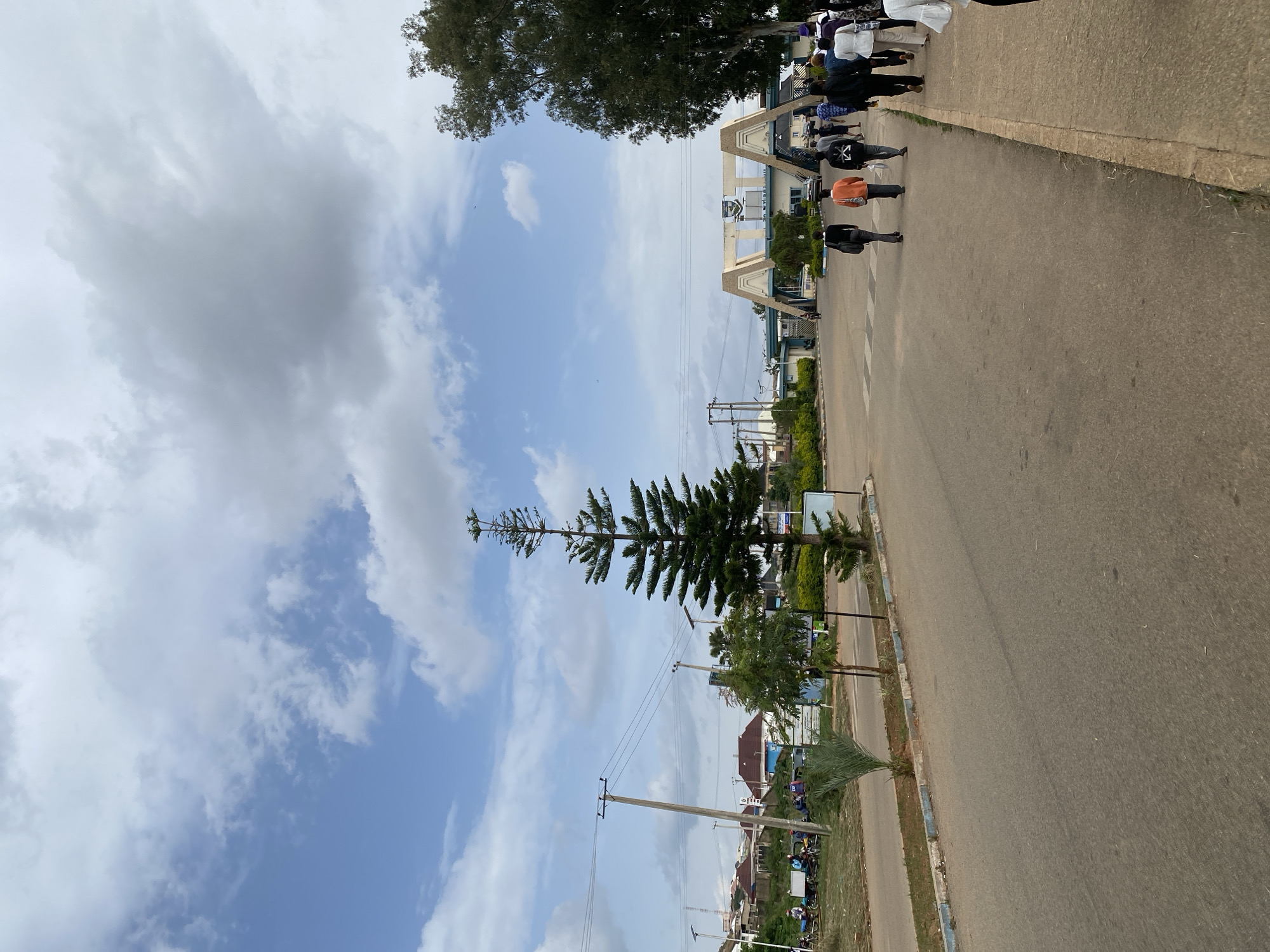 The image depicts a daytime scene along a paved road in Jos, Nigeria. A group of people are walking on the sidewalk beside the road, moving away from the camera. They are dressed in casual clothing, including shirts and trousers. Behind them, a large, arched gateway or entrance structure is visible, with buildings and trees in the background. The sky is partly cloudy with a mix of white and gray clouds, suggesting fair weather. A tall, conical evergreen tree stands prominently on the left side of the frame, reaching towards the sky. Further down the road, utility poles and power lines are present, indicating an urban or semi-urban setting. Several residential buildings with red roofs and some motorcycles are also visible in the distance. The overall atmosphere appears to be neutral, with no strong emotional cues conveyed. The perspective is from a slightly elevated angle, looking down the road and across the sidewalk.