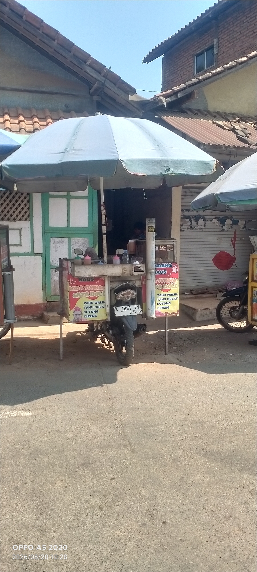 In Bugel, Indonesia, a street vendor operates a food stall from a small, mobile cart. The cart is covered by a large, light green umbrella, providing shade. Behind the cart is a building with a light-colored facade and a corrugated metal roof. Adjacent to the building are closed metal shutters, one of which has a large red lipstick imprint. A motorcycle is parked in front of the cart, its license plate clearly visible and reading "K 4891 ZV". The cart displays signs with Indonesian text, advertising food items like "TAHU WALIK" and "SOTONG". A person is visible sitting inside the stall. The ground is asphalt, with a distinct shadow cast across it, indicating it is daytime with bright sunlight. The scene appears to be a typical street-side business in a moderately populated area.