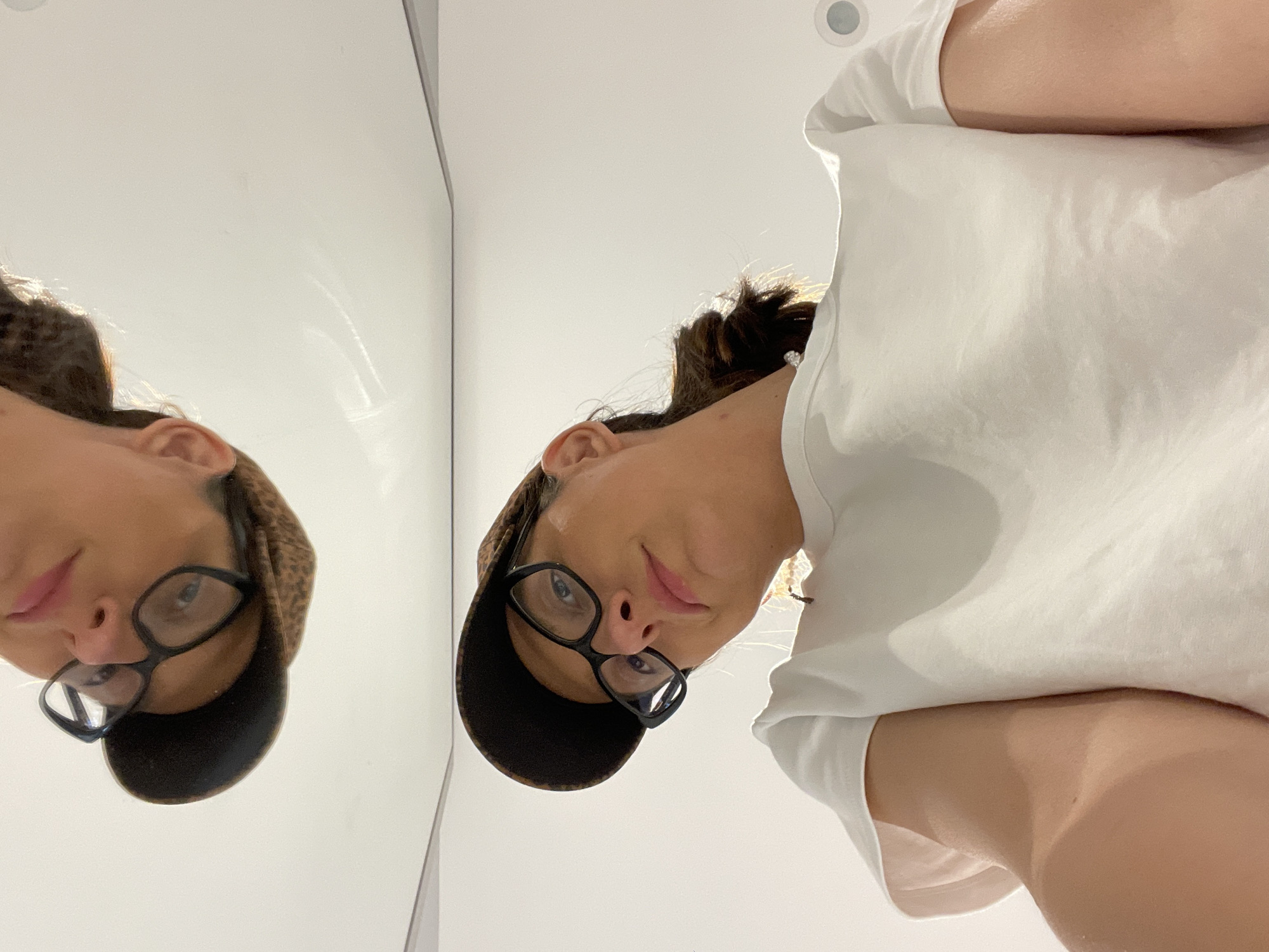 The image presents a selfie-style shot taken from a low angle looking up at a person wearing glasses and a cap. The person is wearing a white T-shirt. To the left is a mirror reflecting the individual's face and part of their cap. The background is plain and white. There is no information to determine if the image was taken in Sofia, Bulgaria or any other place.