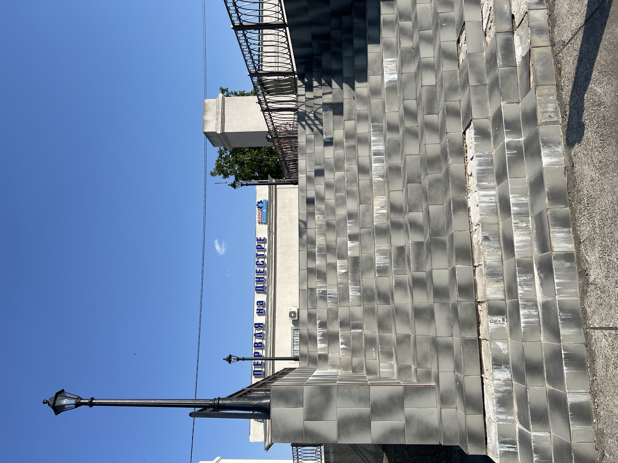 The image depicts a wide outdoor staircase made of grey tiles leading up to a building. The building has blue Cyrillic lettering that reads "ПЕРВАЯ на ДНЕСТРЕ", which translates to "First on the Dniester". A lamppost stands to the left of the stairs, and a black metal railing runs along the side. The sky above is clear and blue. The scene appears to be located in Korzhevo, Moldova. The condition of the staircase shows some wear and tear, with missing and damaged tiles in places, and white streaks running down some of the steps, possibly from water damage. There are no people visible in the image.