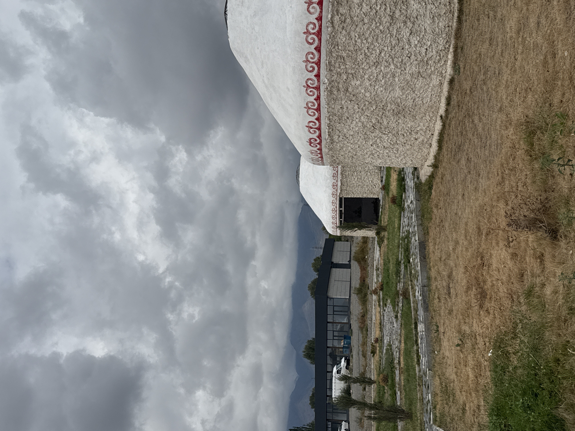 The image captures a scene in Balykchy, Kyrgyzstan, featuring traditional yurts and a modern building under a cloudy sky. In the foreground, dry, brown grass covers the ground, with patches of green grass on the left. A paved pathway with stone borders winds through the area.
The focal point of the image includes two large, white yurts with decorative red patterns around their upper sections. The yurt on the right is more prominent, its textured, stone-like exterior clearly visible. The yurt on the left is partially obscured by the one on the right, revealing an open doorway.
In the middle ground, a modern, rectangular building with large glass windows and a dark-colored roof is situated. Several vehicles, including a white van and a white car, are parked in front of the building. Green bushes and trees are scattered throughout the landscape.
The background is dominated by a mountain range, its peaks softened by the overcast sky. The sky itself is filled with dense, gray clouds, suggesting a cloudy or potentially rainy day. The overall impression is one of a blend of traditional and contemporary elements within a natural, mountainous setting in Balykchy.