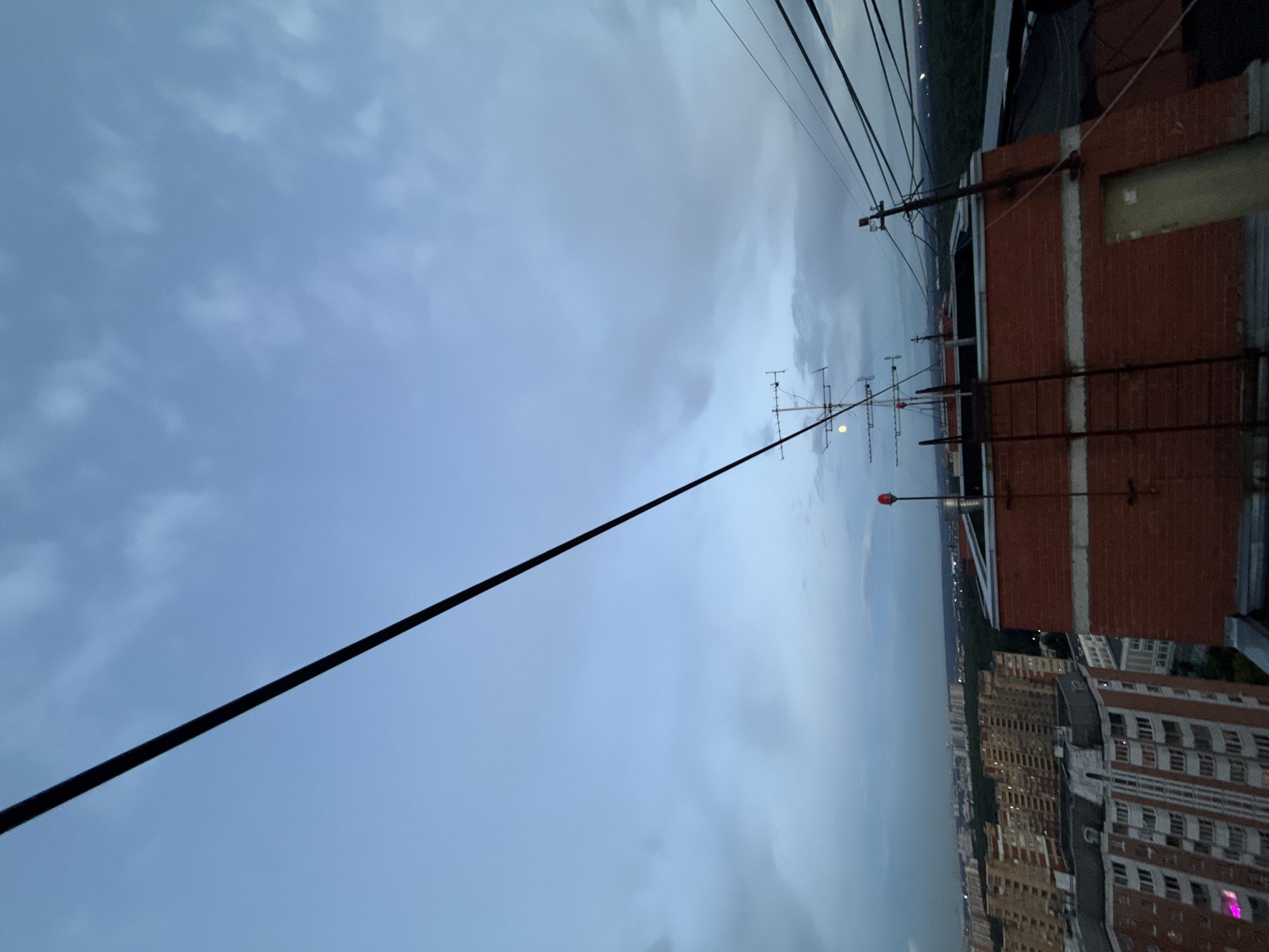The image is a high-angle, vertically oriented shot of a cityscape under a cloudy sky, likely taken at dusk or early evening. The foreground is dominated by the roof of a brick building, with a thick black cable running diagonally across the frame. Other objects on the roof include a TV antenna and what appears to be a red warning light on a pole.
In the mid-ground and background, a dense urban landscape unfolds, characterized by numerous multi-story buildings of varying architectural styles and colors. Some buildings are illuminated, suggesting the city is waking up to the evening hours. The sky is overcast with gray clouds, hinting at potential rain or an impending storm.
The overall scene suggests a view from a high vantage point within a city, possibly a residential or commercial area in Khimki, Russia, given the location context. There is no visible text in the image. The weather appears to be unsettled, and the time of day is transitional, moving from daylight to night. The mood is somewhat somber due to the cloudy skies and the urban sprawl.