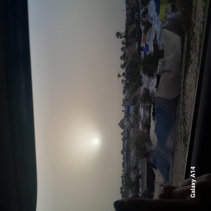 An outdoor landscape shows many tents on a dusty ground under a pale sky with the sun low in the sky, suggesting dawn or dusk. There are palm trees scattered throughout. The view seems to be from inside a vehicle as part of a car window is visible. The camera is a Galaxy A14.