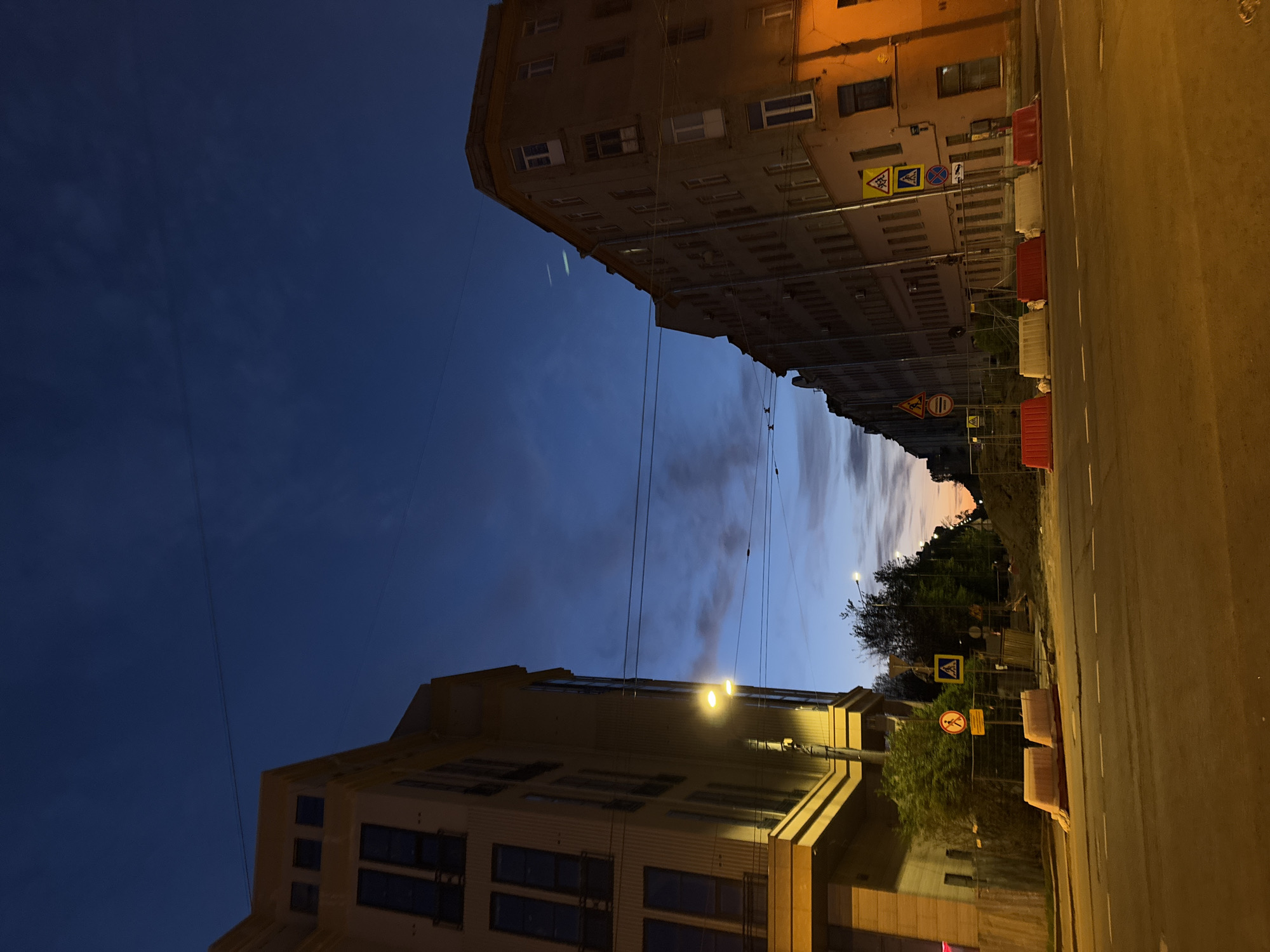 Captured at night in Saint Petersburg, Russia, the image showcases a street scene framed by buildings on either side. The sky above transitions from a deep blue to lighter hues near the horizon. Overhead wires crisscross between the buildings. The street is lit by streetlights, and temporary construction barriers line the side, accompanied by various traffic signs, including pedestrian crossing warnings. No people are visible in the frame.