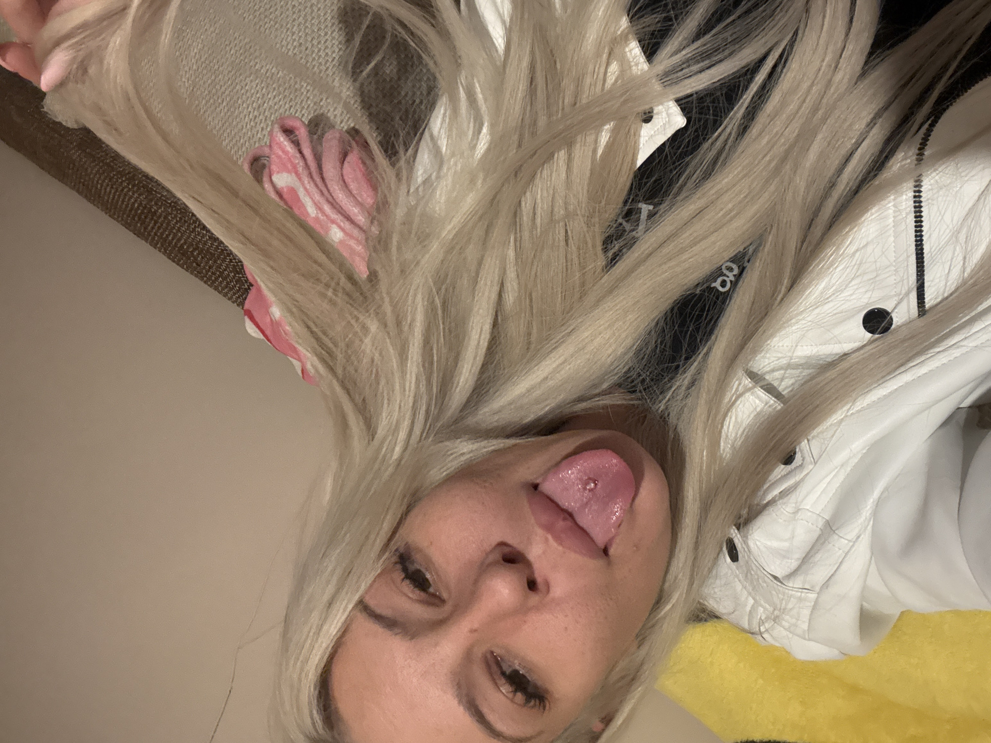 A blonde woman with a tongue piercing is visible in the image. She is wearing a white jacket with a black zipper and black accents. A pink object is visible behind her hair. The background includes a beige wall or surface and a glimpse of yellow material. The location is not discernible from the image; therefore, it is impossible to identify the location in Plovdiv, Bulgaria.
