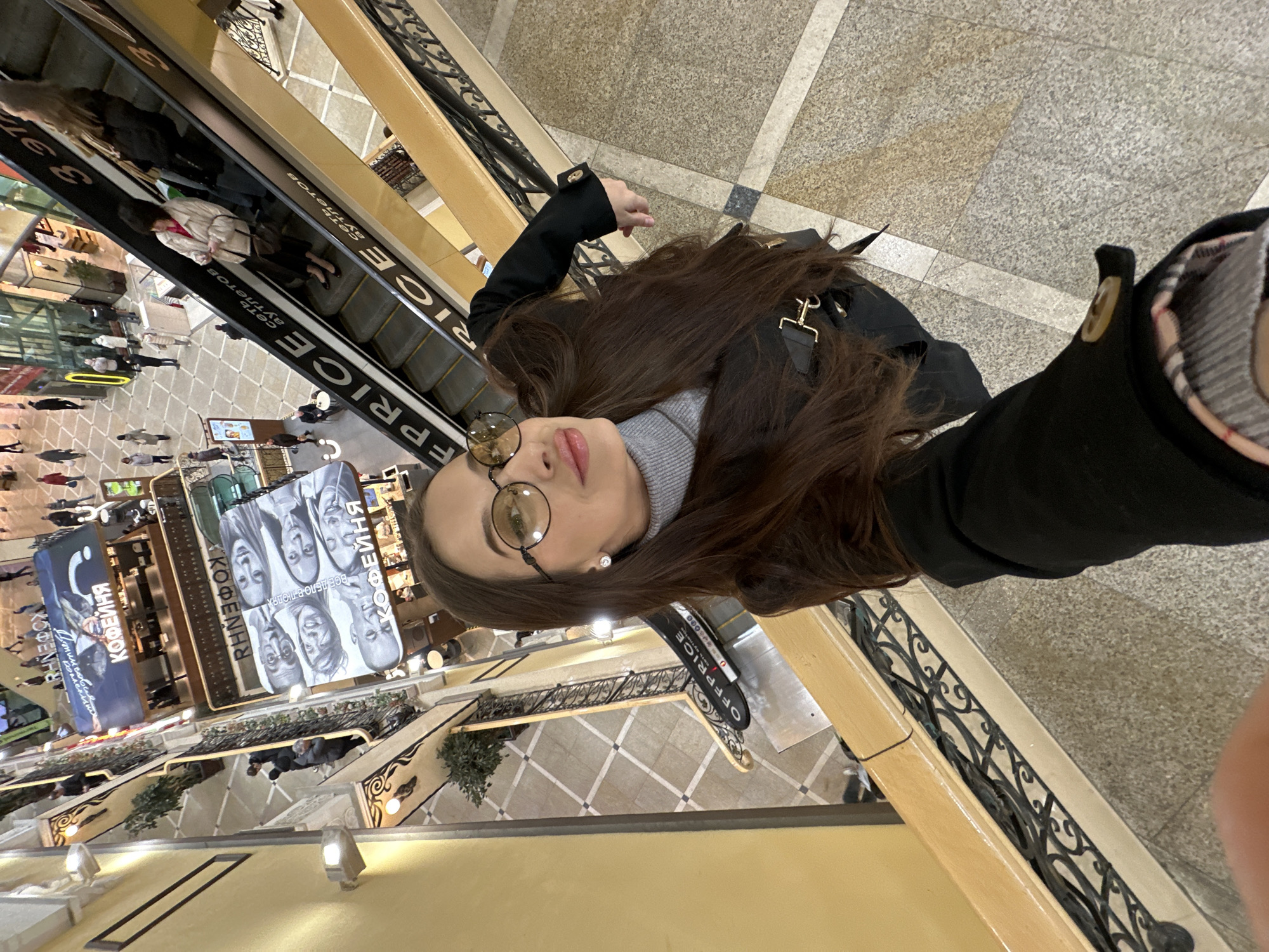 The image is a selfie taken by a young woman from an upper floor of a multi-level shopping mall or commercial complex in Yekaterinburg, Russia. The main subject, with long dark hair, round glasses, a grey turtleneck, and a black coat, looks towards the camera with a neutral expression, holding the phone from an elevated position.
Below her, an escalator is visible, displaying the text "OFFPRICE" and "СЕТЬ АУТЛЕТОВ" (Outlet Network), with several blurred figures riding it. The mall features light-colored tiled floors, some with intricate patterns, and decorative black metal railings along the balconies. On the lower floors, numerous shoppers can be seen, along with retail storefronts and displays.
Prominently displayed on the lower level are large banners. One banner features several black-and-white portraits with the Russian text "ВСЕ ДЕЛО В ЛЮДЯХ" (It's all about people) and "КОФЕЙНЯ" (Coffee Shop). Another banner also advertises "КОФЕЙНЯ". The interior is brightly lit, indicating a vibrant commercial environment. The scene captures a moment of leisure or shopping in a modern Russian mall.