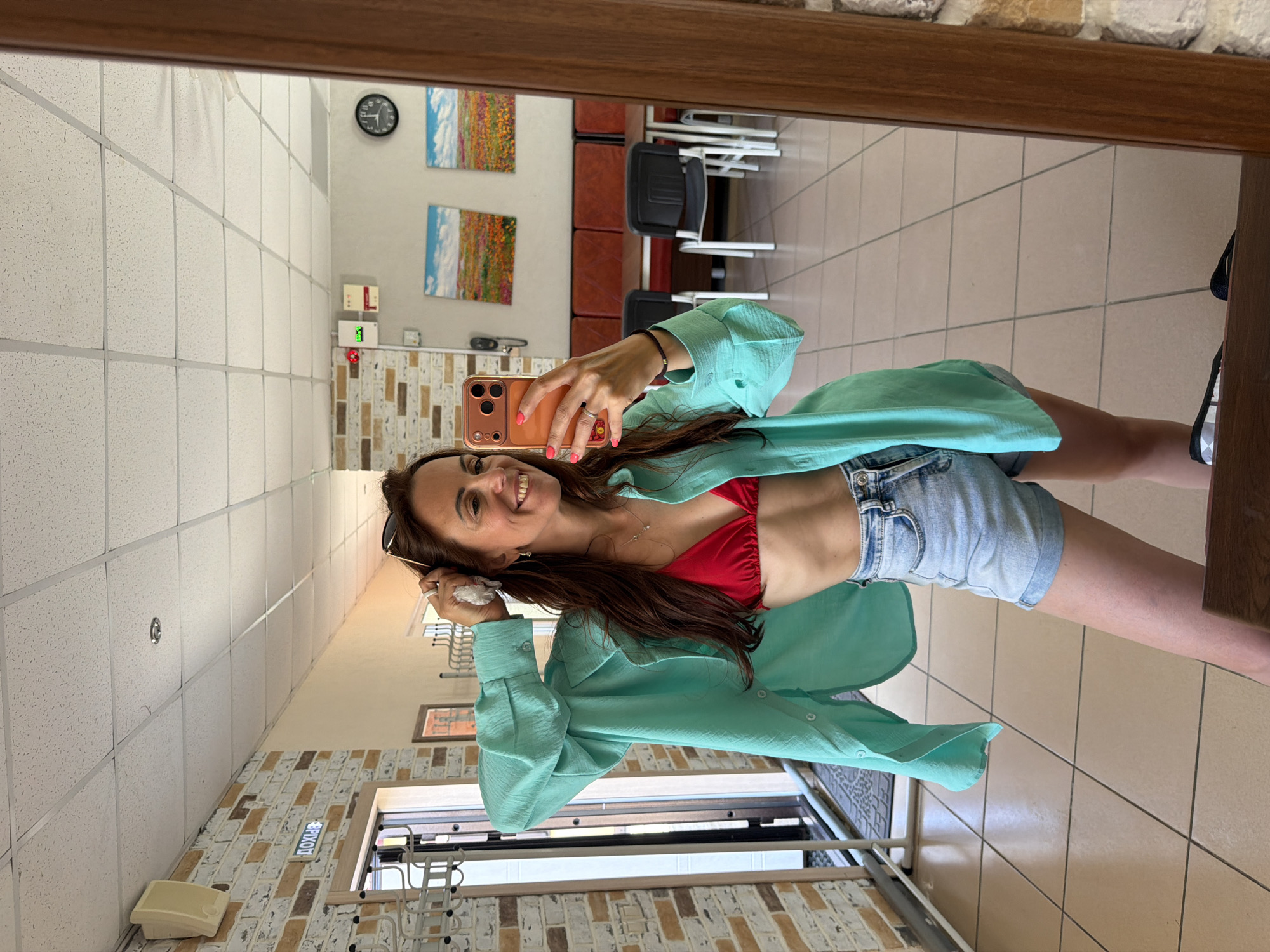 A woman with long, dark brown hair, wearing a red bikini top, denim shorts, and an open, light blue oversized shirt, takes a selfie in a mirror. She is smiling and holding her phone in her right hand, with her left hand touching her hair. She is standing in what appears to be an indoor common area, possibly a cafe or waiting room, with tiled floors and brick walls. In the background, through the reflection in the mirror, several tables and chairs are visible, along with artwork on the wall and a clock. A sign with Cyrillic text, "ДОХОД", is visible on the brick wall. The overall scene suggests a casual, indoor environment. There is no information in the image to suggest the location is in Birobidzhan, Russia.