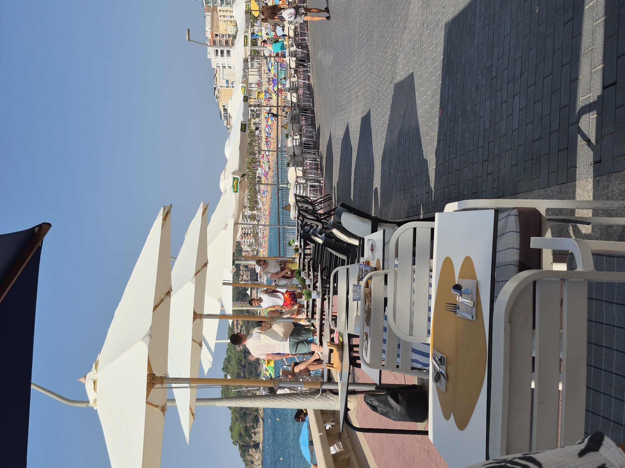 This image depicts a sunny outdoor dining area in Lloret de Mar, Spain, likely situated along the promenade overlooking the beach. The scene is dominated by several rows of outdoor tables and chairs, many covered by large white umbrellas. The tables are set with placemats and cutlery, indicating a restaurant or cafe.
In the background, the sandy beach is visible, crowded with beachgoers and adorned with numerous colorful umbrellas. The turquoise water of the Mediterranean Sea stretches out beyond the beach, with a coastline featuring hills and buildings that form the cityscape of Lloret de Mar.
Several people are present in the scene. In the foreground, a table is set for dining. Further back, individuals are seen walking along the promenade, some are seated at tables, and others are standing. The overall atmosphere is one of a bustling tourist destination on a clear, warm day. The paving stones of the promenade are clearly visible, as is a low wall separating the dining area from the beach. Shadows from the umbrellas and structures are cast across the promenade.