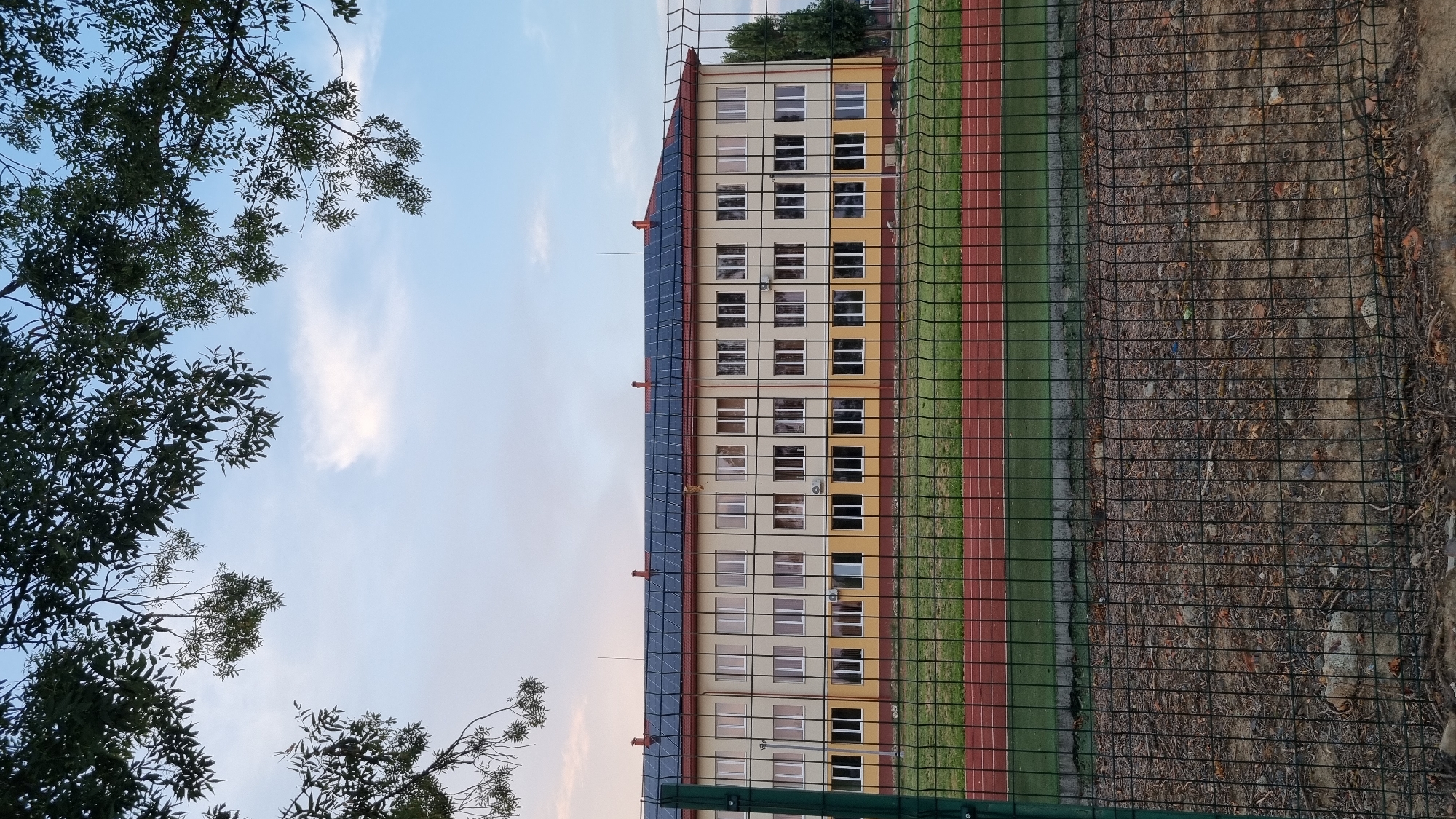 The image shows a school building with solar panels on its roof. The building is a two-story structure with many windows. A fenced sports field, including a running track, is in front of the school. The sky is partly cloudy. The location is the school in Berehove, Ukraine.