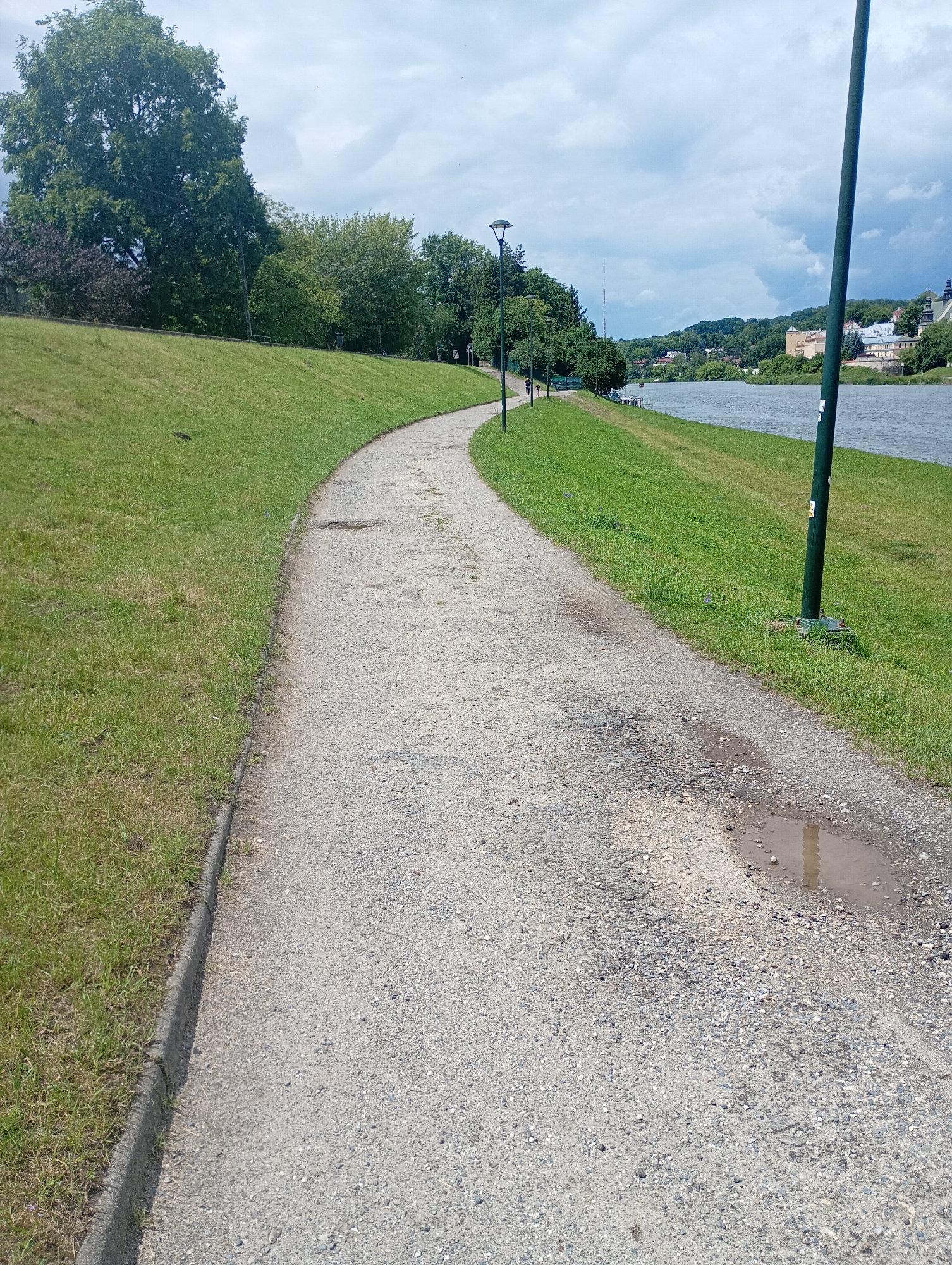 An outdoor, eye-level, vertical-format, daylight shot captures a paved walking path curving along the bank of a wide, calm river. The path is made of a light-colored gravelly material, and is in somewhat disrepair, with visible potholes and a puddle of water. To the left of the path is a gently sloping, well-maintained grassy hill dotted with a few small dark objects, possibly animals. Several trees are visible on the top of the hill.
To the right of the path, the river stretches out, with a few small boats docked on the far bank. Across the river, a picturesque town with buildings of various colors and architectural styles is nestled amongst green, rolling hills. The sky above is filled with dramatic, white and gray clouds. Two lampposts are visible along the path, and a few distant figures can be seen walking or cycling. The overall scene evokes a sense of peaceful recreation and natural beauty within an urban setting. This scene is located in Krakow, Poland, along the Vistula River.