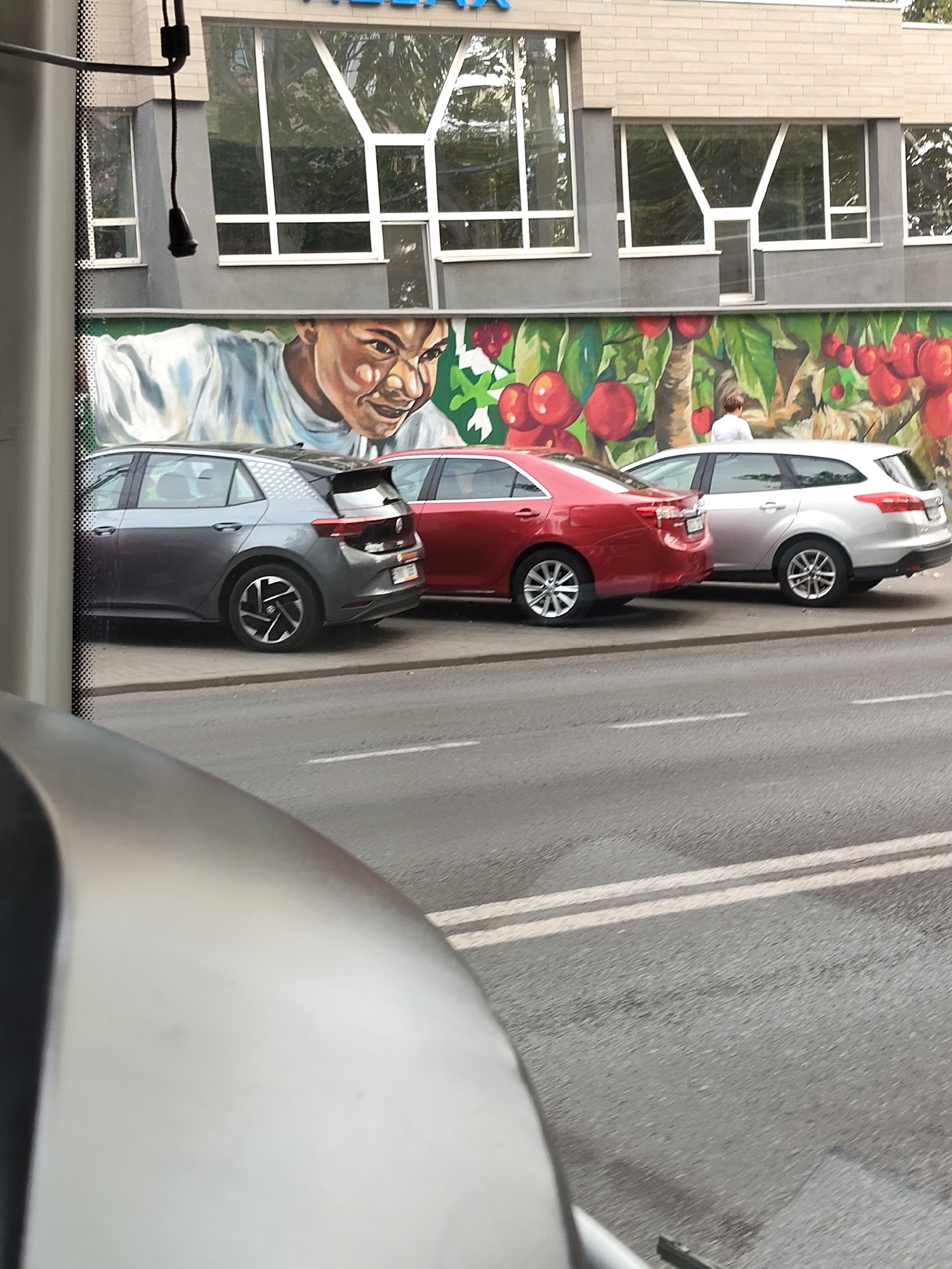 The image shows a mural of a person's face and fruit on the side of a building. Three cars are parked in front of the mural. The cars are a gray hatchback, a red sedan, and a silver station wagon. The scene appears to be on a street in a city. The cars are parked parallel to the curb. The mural is vibrant and colorful, with a close-up of a person's face and large red fruits. The building behind the mural has large windows.