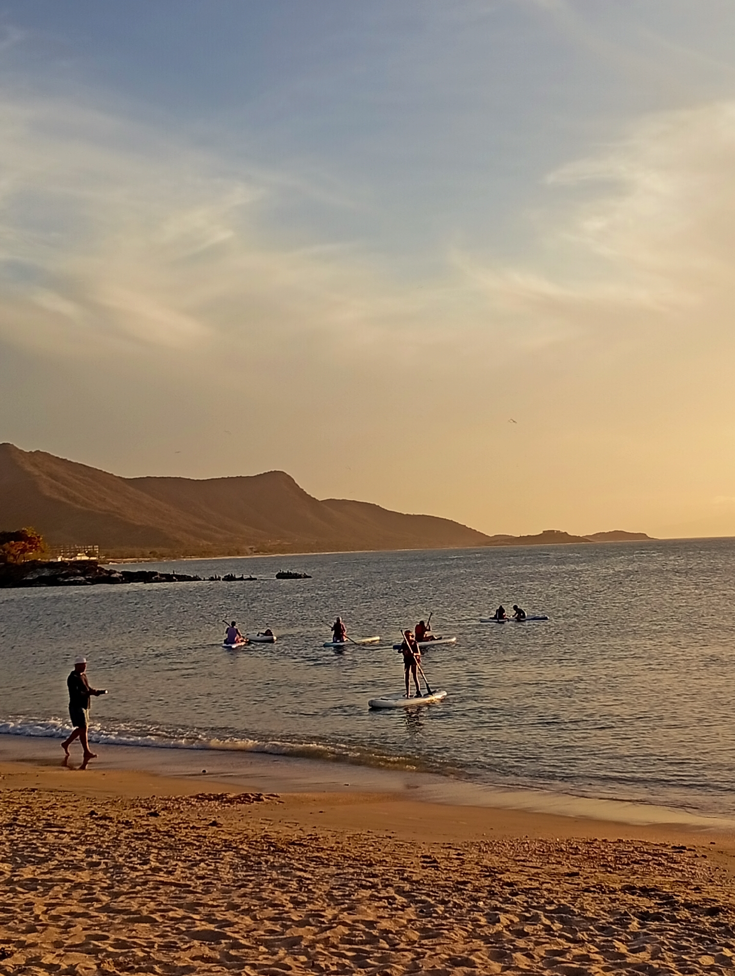 The photograph shows a beach scene at sunset in Juan Griego, Venezuela. In the foreground, sandy beach stretches into the distance, with a man walking barefoot along the shore. He is wearing dark clothing and a light-colored hat. In the water, several people are paddleboarding, scattered at varying distances from the shore. Mountains form a backdrop in the distance, silhouetted against the sky. The sky is a mix of light blue and golden hues.