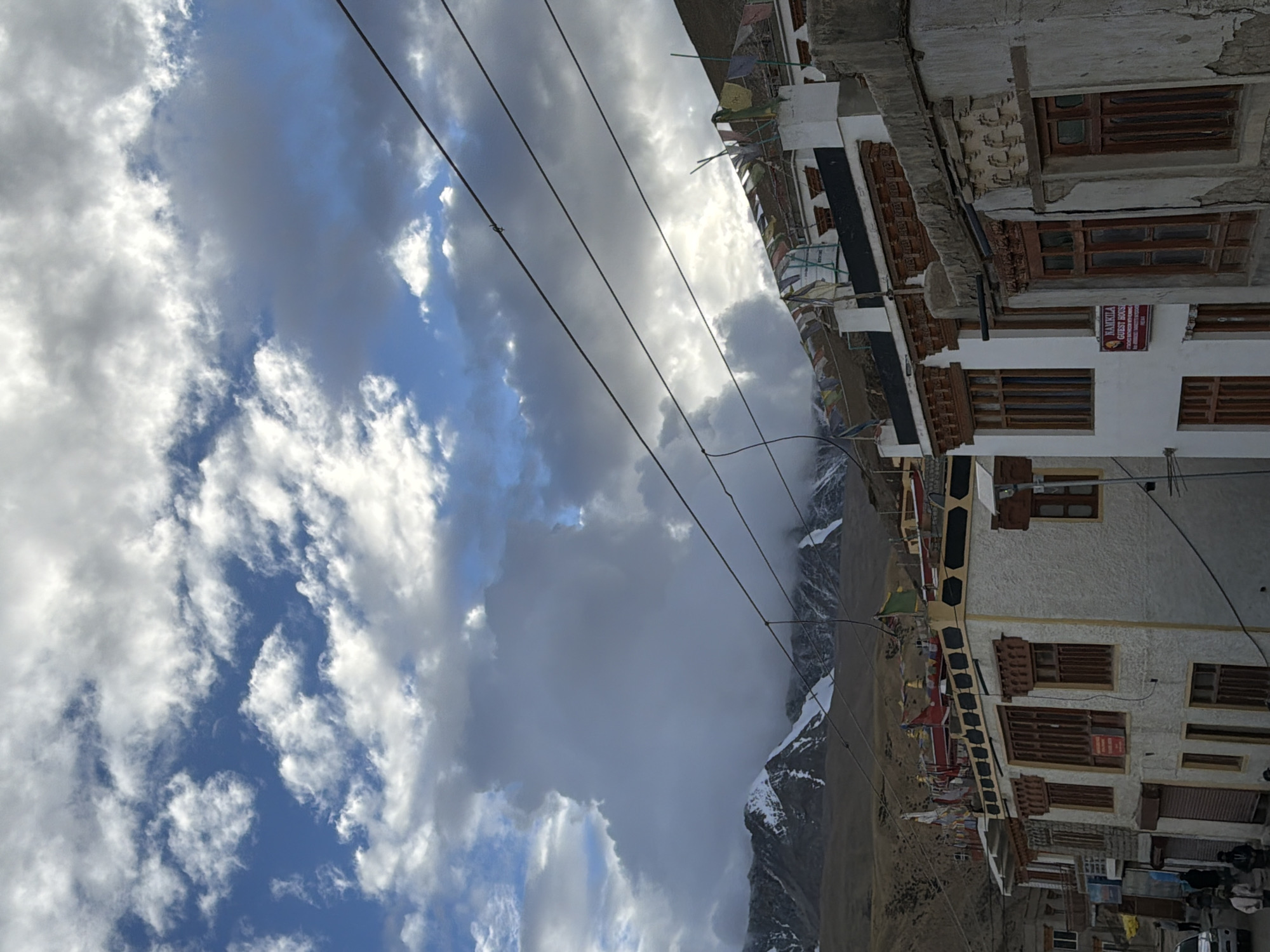 The image depicts a street scene in Karzok, India, characterized by traditional architecture and mountainous terrain. In the foreground, several multi-story buildings with white stucco exteriors and wooden window frames line the street. Prayer flags are strung across the rooftops of these buildings. A sign on one building reads "NAMKILA GUEST HOUSE". Further down the street, glimpses of other buildings and a vehicle can be seen. In the background, snow-capped mountains rise, partially obscured by dramatic clouds. Utility wires are visible stretching across the sky, adding a modern element to the otherwise traditional setting. The sky itself is a mix of blue patches and heavy, dark clouds, suggesting dynamic weather conditions.