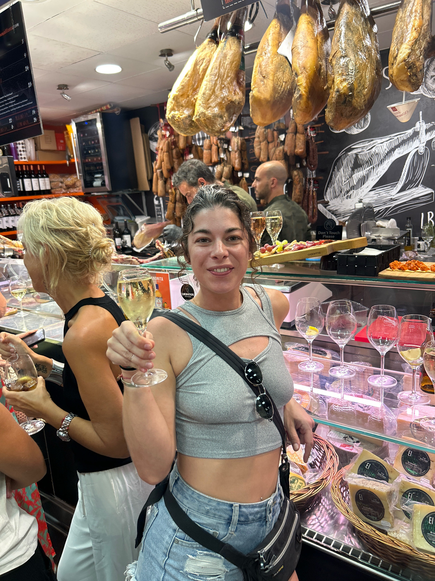 In a market stall in Palma de Mallorca, Spain, a woman with curly dark hair is holding up a glass of white wine, smiling at the camera. She is wearing a grey crop top and denim shorts, with a black fanny pack slung across her body. Behind her, other people are visible, including a woman with blonde hair and a man with a beard. The stall is filled with hanging meats and shelves of cheese and wine. The overall atmosphere appears to be lively and social.