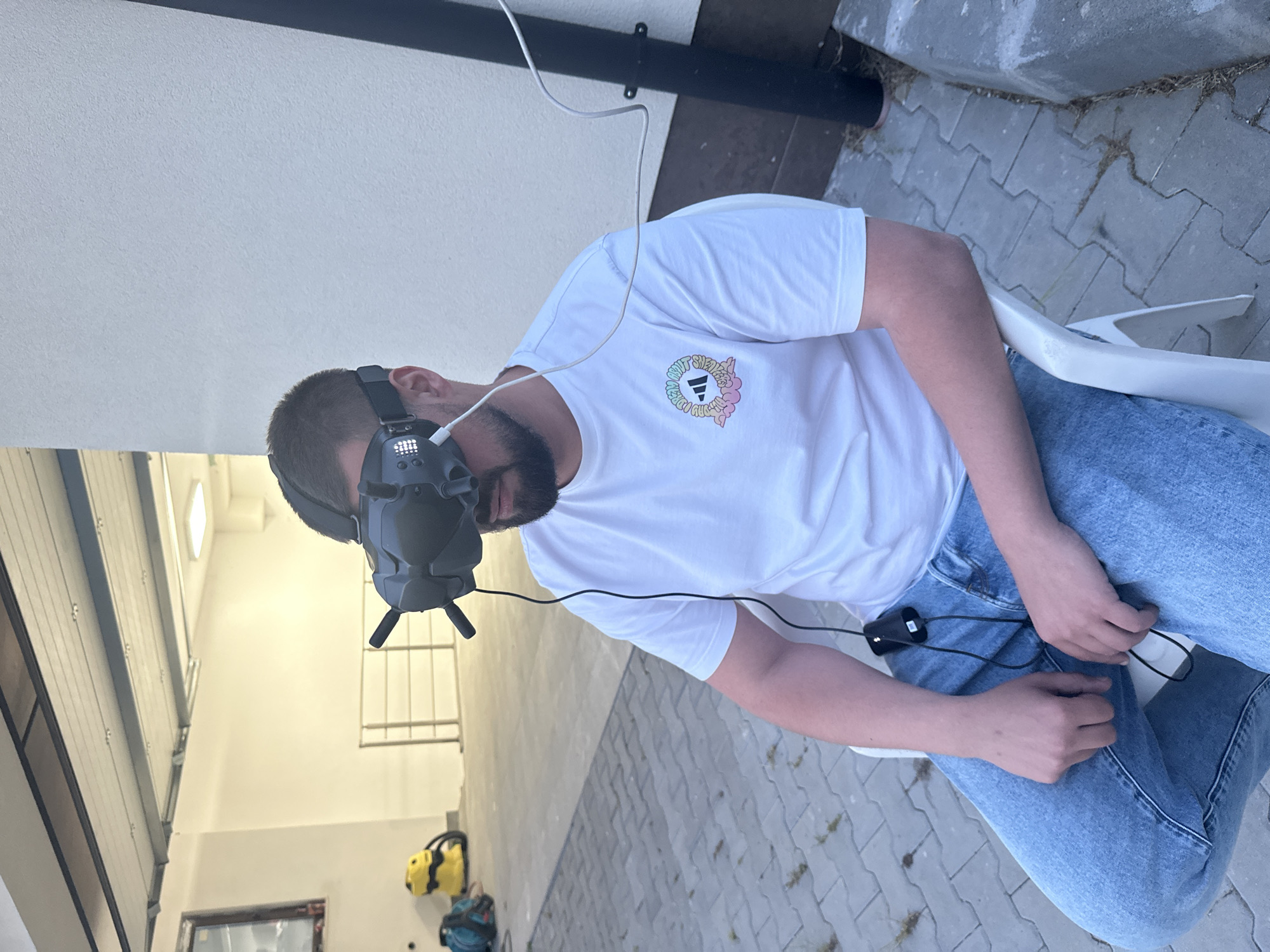 A man with short hair and a beard is wearing a grey VR headset and a white t-shirt with a round logo on the left side. He is sitting on a white plastic chair, wearing blue jeans. A black wire connects the VR headset to a device on his lap. The background shows a white wall and a brick paved ground. Inside a garage, there is a blue backpack, a yellow chainsaw, and a window. Based on visual cues of architectural style and the types of materials used, it is likely that the location is in Plovdiv, Bulgaria.