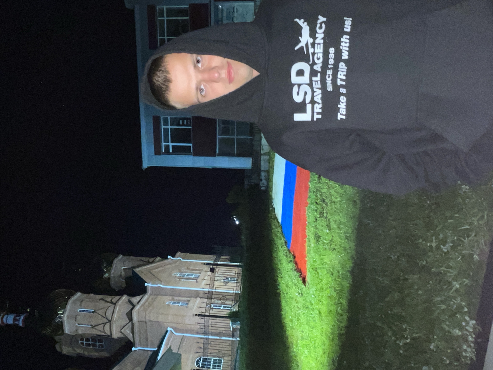 The image, presented rotated 90 degrees clockwise, depicts a young person posing at night in what appears to be Красные Баки, Russia. In the foreground, a person, likely male, faces the camera with a neutral to slightly smirking expression. They wear a dark hooded sweatshirt with the hood pulled up, framing their face and revealing short, dark hair.
Prominently displayed on the sweatshirt in white text is "LSD TRAVEL AGENCY" with a small airplane graphic, followed by "SINCE 1938" and "Take a TRIP with us!".
The setting is outdoors at night, with the background plunged into darkness. Artificial light illuminates the immediate foreground and parts of the person. To the person's right, a large Russian flag, featuring horizontal white, blue, and red stripes, is laid out on bright green grass. In the deeper background, two buildings are discernible: one appears to be a multi-story, architecturally elaborate structure with arched windows, possibly a church or historic building, while another, simpler building with rectangular windows is partially visible. The dark sky confirms the nighttime setting.
