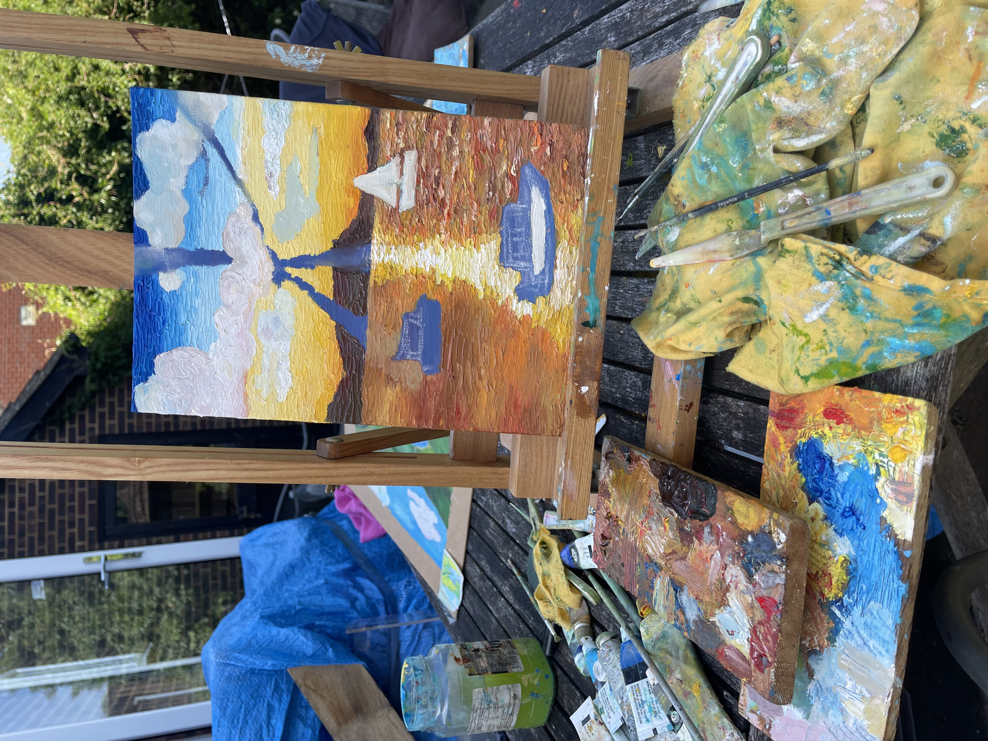 The image depicts an outdoor art setup on a weathered wooden table. A partially completed oil painting of a sunset with boats and reflections sits on a wooden easel. To the right of the easel is a yellow cloth stained with paint, along with several paintbrushes and palette knives. Two wooden paint palettes covered in various colors of oil paint are positioned in the foreground, surrounded by tubes of paint. Underneath the table, a blue tarp is visible, along with a glass jar with a green lid containing a blue liquid. Based on the building visible in the background, the location can be determined to be Maidstone, United Kingdom. No people are visible in the image.