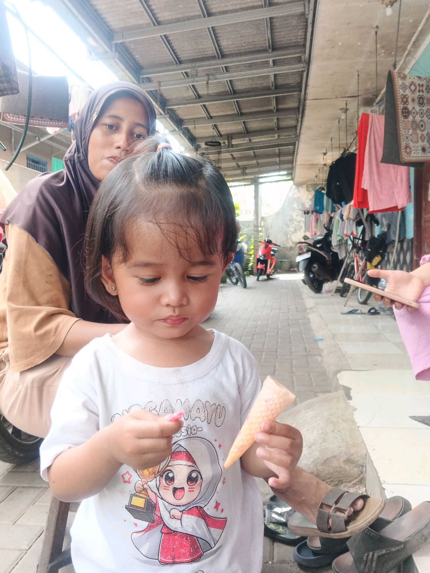 The image depicts a young child, likely a girl, in the foreground, holding an empty ice cream cone. She is wearing a white t-shirt with a cartoon graphic. To her left and slightly behind her, a woman wearing a hijab and a brown and tan garment is partially visible. The background suggests an outdoor, possibly residential area with a corrugated metal roof overhead, laundry hanging on lines, and several motorcycles parked. The ground is paved with interlocking bricks. The scene appears to be in Kadu, Indonesia, given the context.