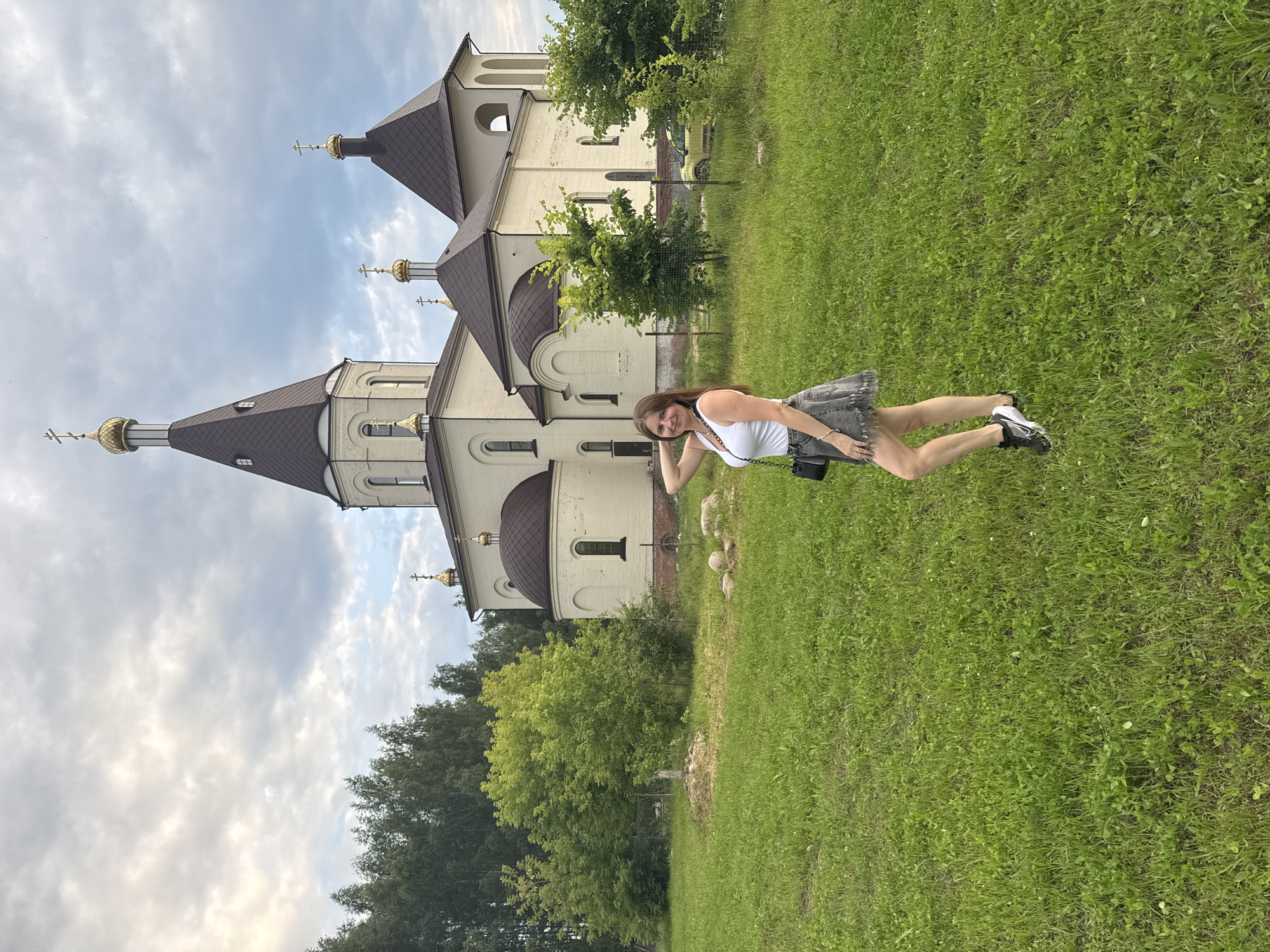 In Konakovo, Russia, a young woman with long brown hair stands in a grassy field in front of a light-colored church with a dark, pointed roof and gold domes topped with crosses. She is wearing a white tank top, a grey denim skirt with a fringed hem, and black and white athletic shoes. She has a black purse with a chain strap slung over her shoulder and her right arm is bent at the elbow with her hand resting on her head. The church is surrounded by trees, and a cloudy sky hangs overhead. A yellow car is parked in the background.