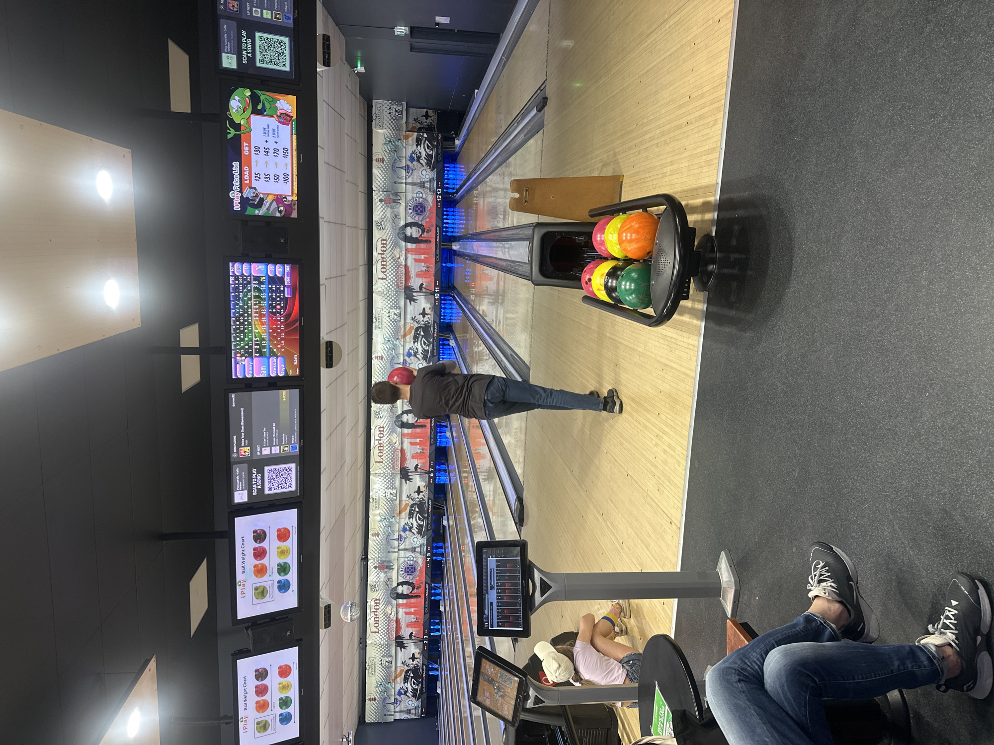 The image shows a bowling alley with multiple lanes. In the foreground, a person is sitting on a chair with their legs crossed, wearing blue jeans and sneakers. To the left, a young girl in a white cap and pink shirt is sitting on the floor facing a digital screen. A bowling ball rack with several colored bowling balls is visible in the middle ground. A man is standing on the right, holding a red bowling ball. In the background, bowling lanes are set up with pins at the end of each lane. Digital screens above the lanes display information, including ball weight charts and current song selections. The bowling alley appears to be located in Coffs Harbour, Australia, based on the "iPlay Coffs Harbour" branding visible on the screens and the "London" and "Wyoming" signage on the backdrop of the lanes, which is common for entertainment venues.