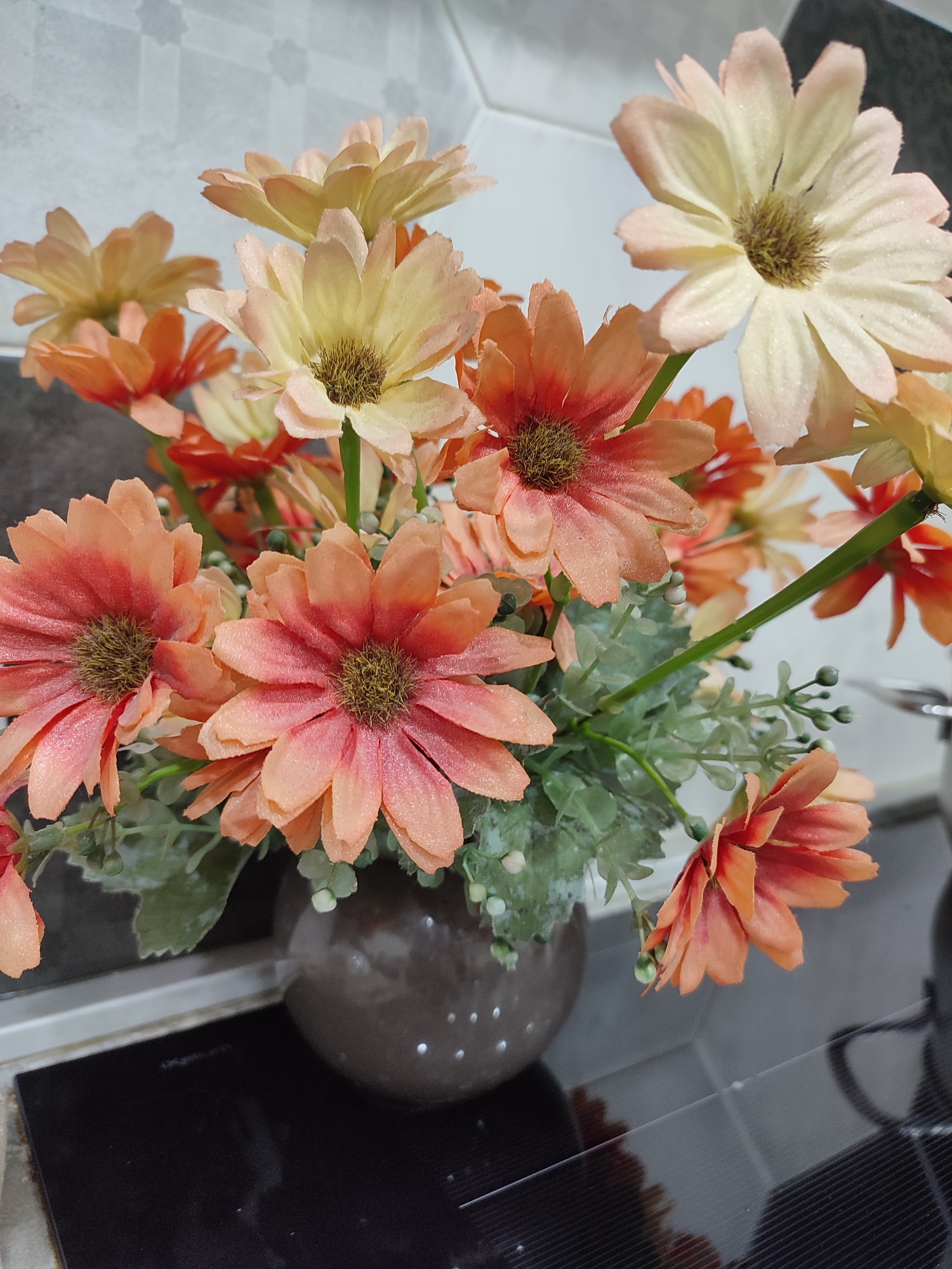 A close-up shot captures a bouquet of artificial flowers in various shades of orange and pale yellow, arranged in a dark brown, round vase. The flowers, resembling daisies, are depicted with textured petals and brown centers, some appearing slightly dewy. Lush green artificial foliage intertwines with the blooms. The arrangement sits on a dark, reflective surface, likely a countertop, with a tiled backsplash visible in the background.