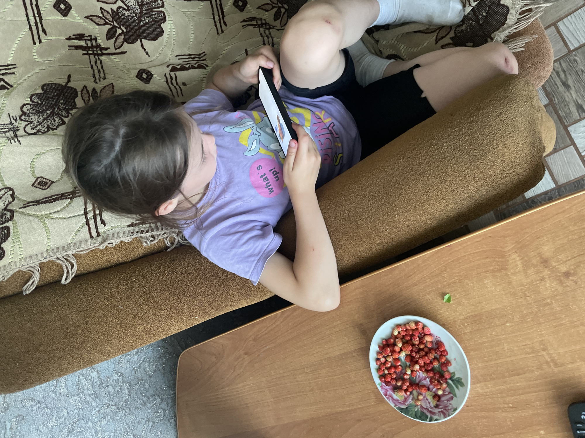 A young girl sits on a sofa, holding a smartphone. She is wearing a purple t-shirt with a cartoon character and text that reads "what's up!". She also wears black shorts and white socks. The sofa is brown and has a patterned blanket on it. To the left of the sofa, on a wooden table, is a plate of wild strawberries and a single green leaf. The flooring is a mix of grey and white tiles. The setting appears to be a residential indoor environment, possibly in Nizhnii Bugrysh, Russia, given the context provided.