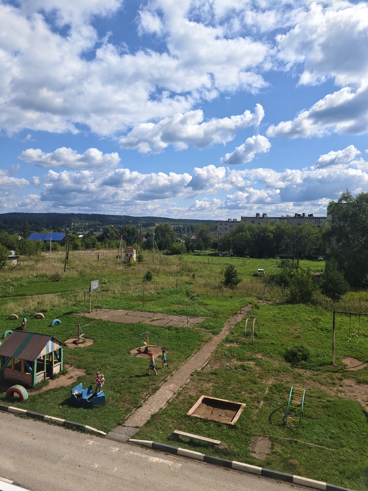 An aerial, eye-level view captures a children's playground and surrounding residential area in Kurashim, Russia, under a bright blue sky dotted with fluffy white clouds. The playground features a small, colorful playhouse, a sandbox, swings, a basketball hoop, and various other play structures. Several children are visible playing on the equipment, including two boys running, a woman holding two children, and another child on the swings. The playground is surrounded by a grassy area, with trees and buildings in the background. A paved road with a sidewalk runs along the bottom of the frame. The scene depicts a typical summer day in a Russian residential neighborhood.