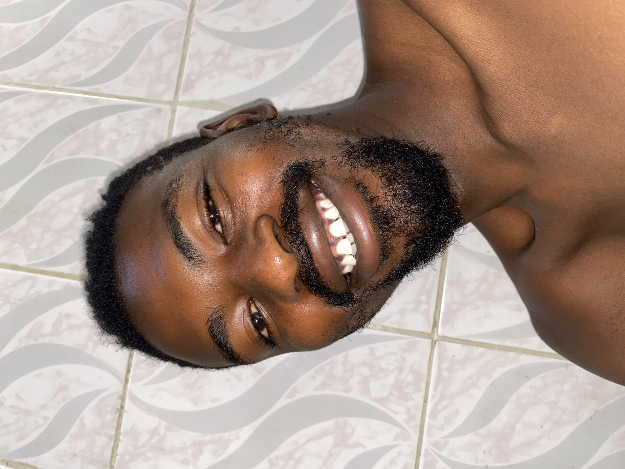 The image captures a close-up of a Black man, likely in Barakin Akawo, Nigeria, looking directly at the camera. He is lying down, with his head positioned towards the lower left and his upper body extending towards the upper right of the frame. The man has a full, dark beard and mustache, and short, curly hair. His skin is dark, and he is smiling broadly, revealing his white teeth and conveying an expression of happiness and cheerfulness.
The background consists of a light-colored tiled surface, possibly a floor, featuring distinct grout lines and a repetitive, light grey wavy pattern. The lighting suggests an indoor setting, and no specific time of day or weather conditions can be determined from the image. There is no visible text.